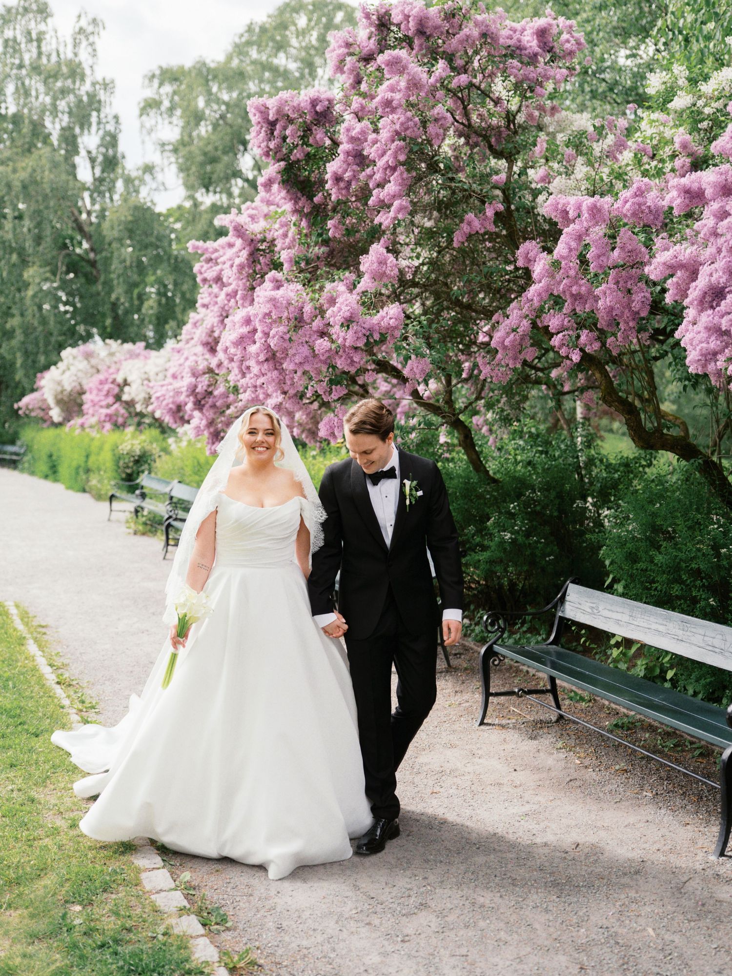 Bride and groom walking hand in hand on a garden path beside blooming purple lilac trees on their wedding day.