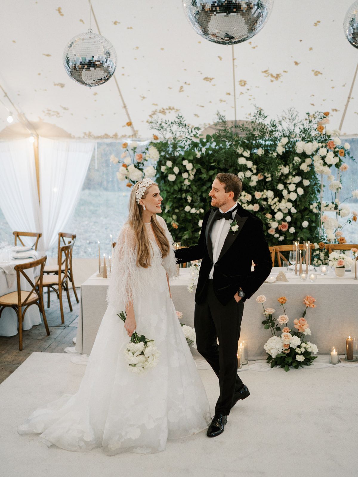 Bride and groom stand beneath disco balls in a floral-decorated tent reception venue at dusk.