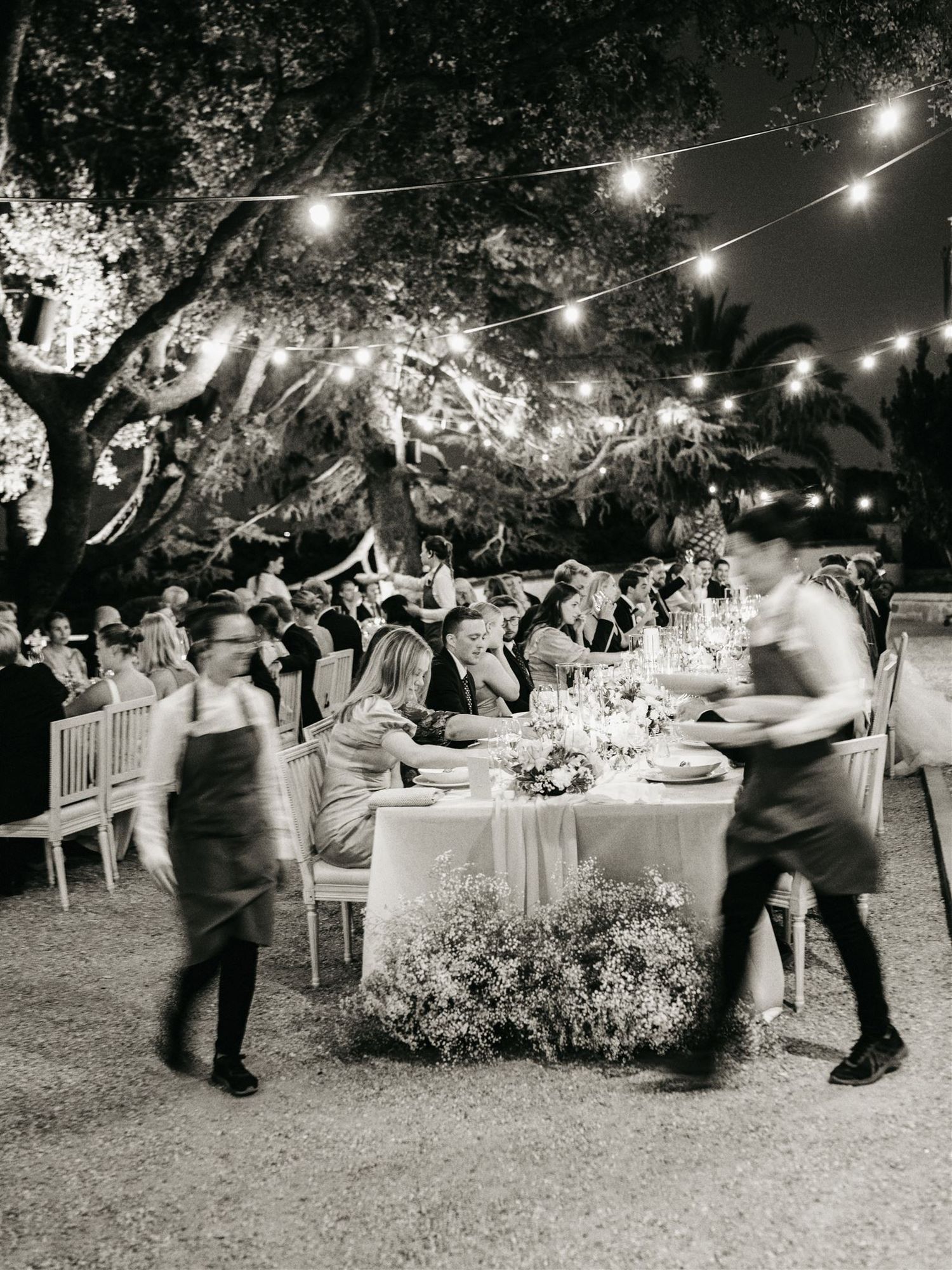Outdoor wedding reception under string lights with waitstaff serving guests at elegant night dinner.
