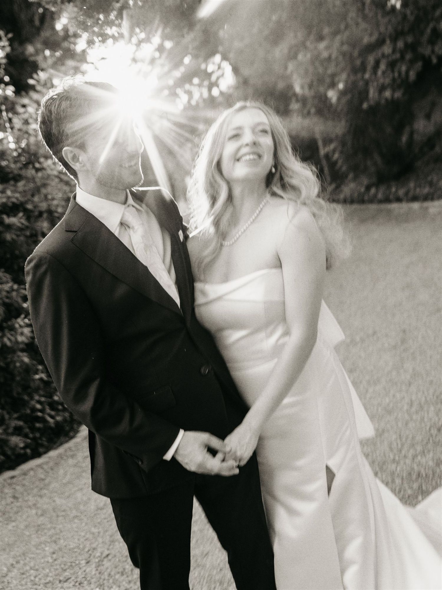Black and white portrait of smiling bride and groom with sun flare in garden setting.