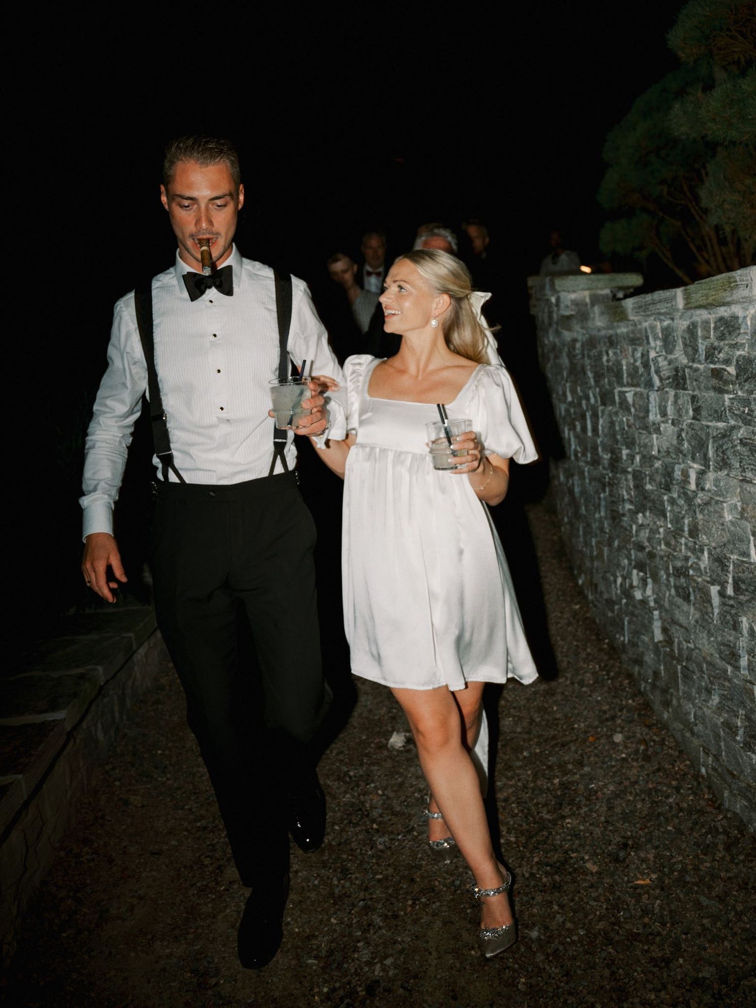 Bride in white mini dress and groom in suspenders walking together at night reception.