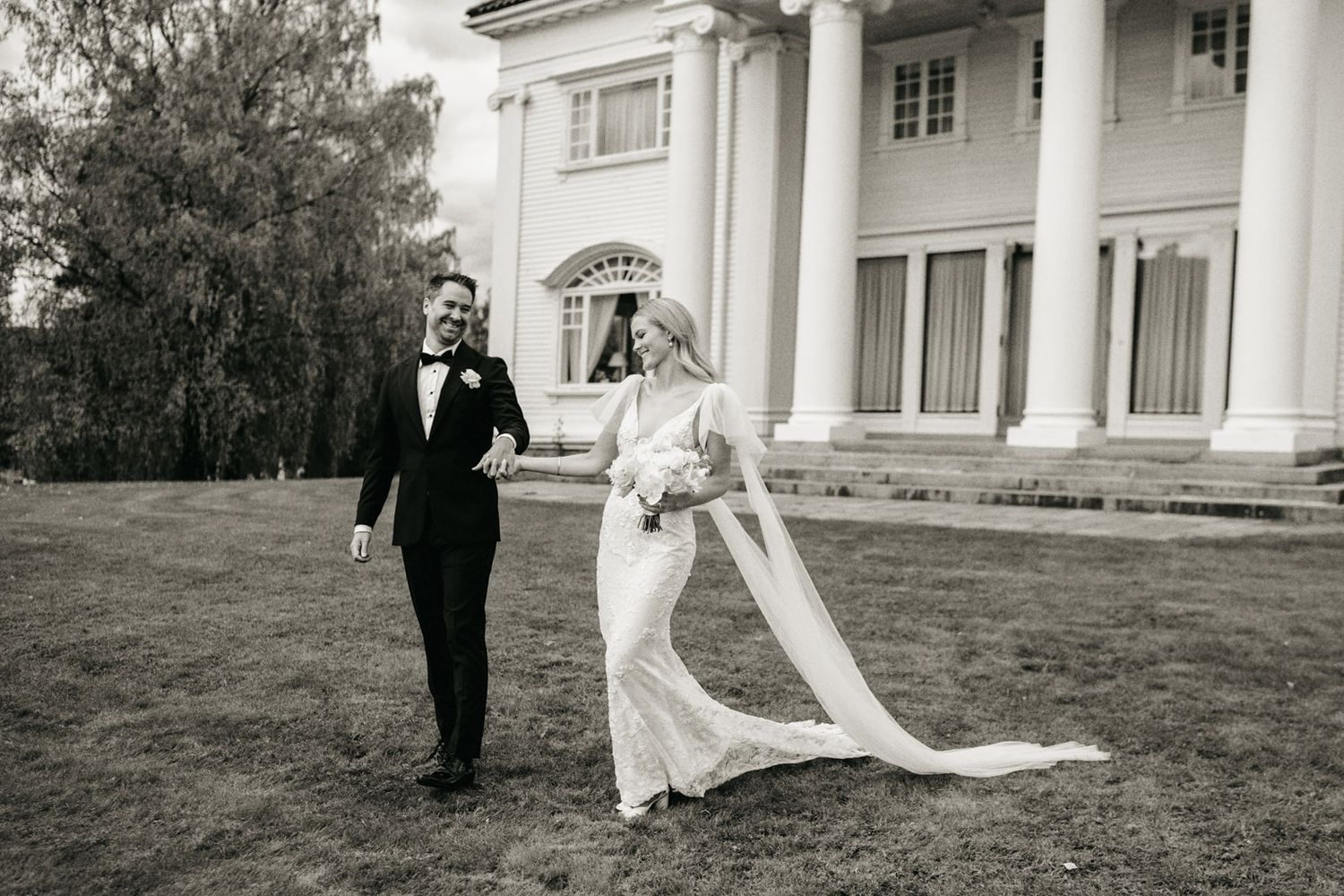 Black and white photo of bride and groom walking hand in hand on lawn before a grand white mansion.