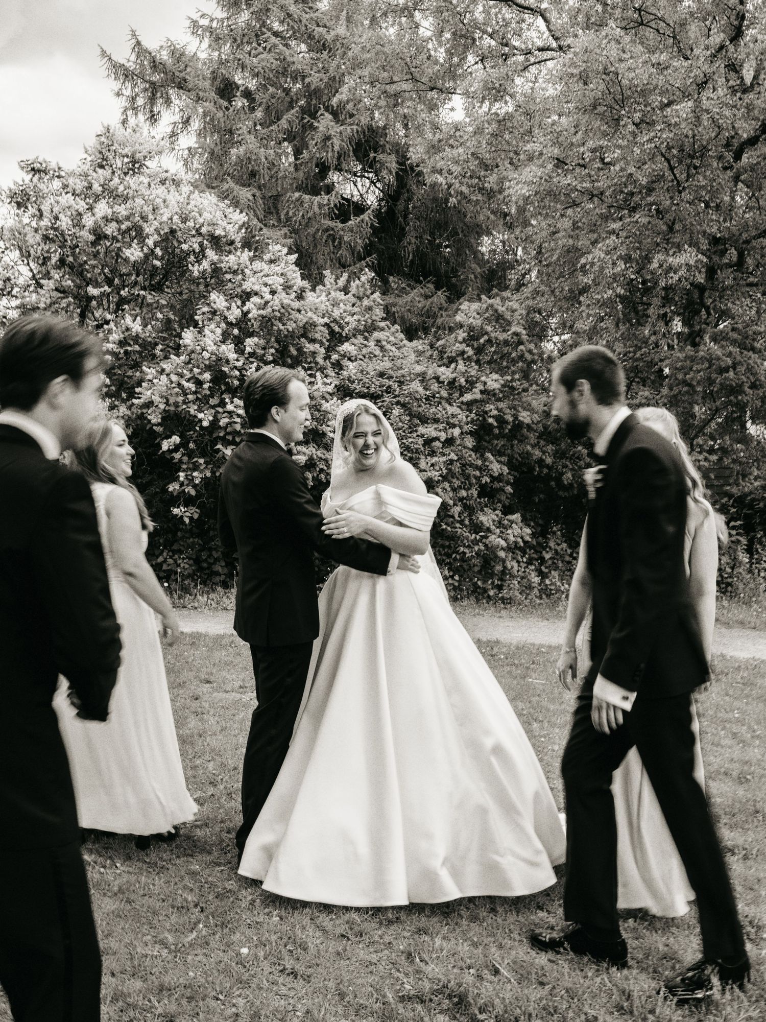 Black and white photo of a bride in a ball gown with groom and wedding party on a lush garden lawn.