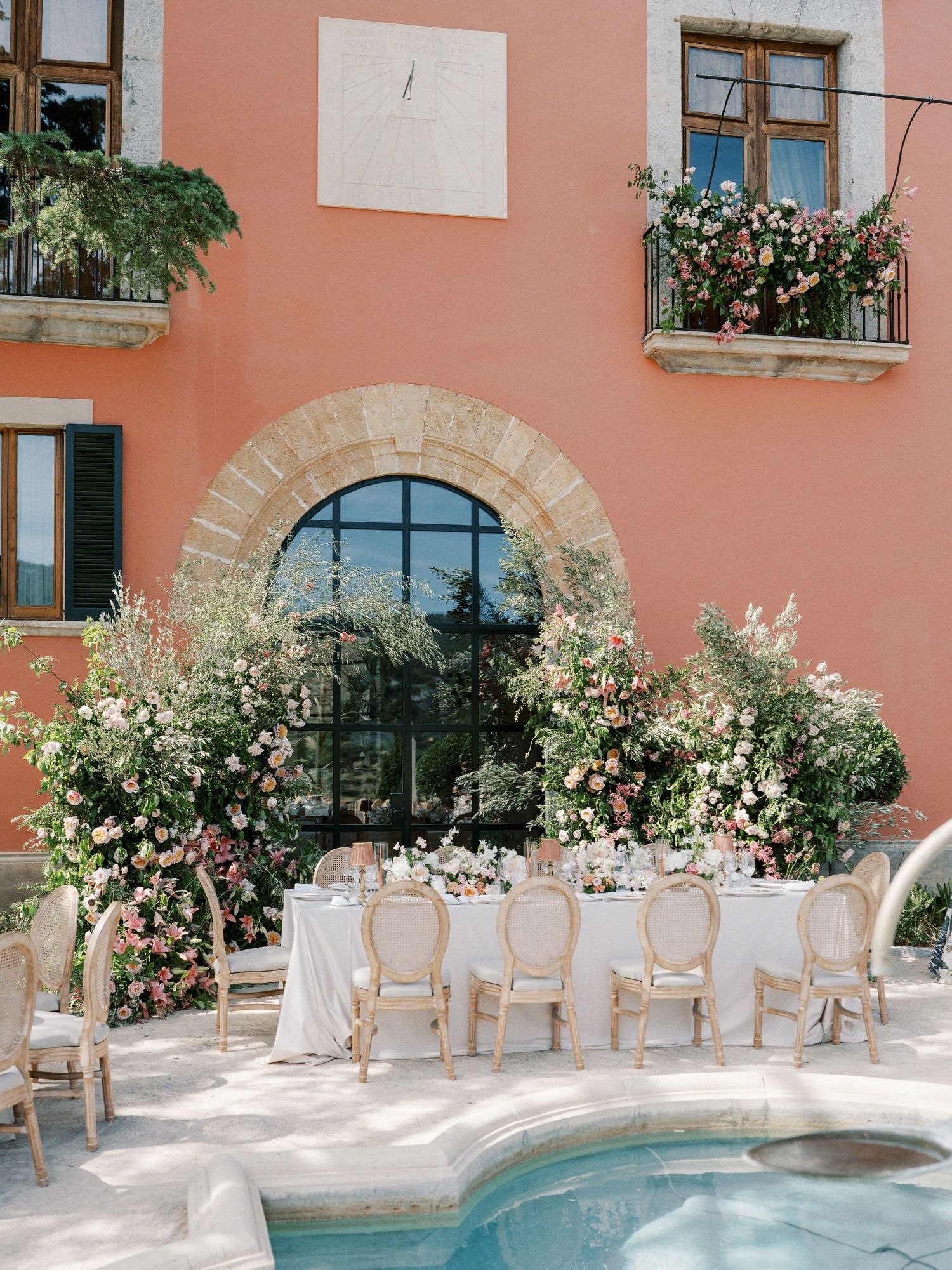 Elegant outdoor wedding reception by a pool at a pink Italian villa, adorned with lush floral arrangements and gold chairs.
