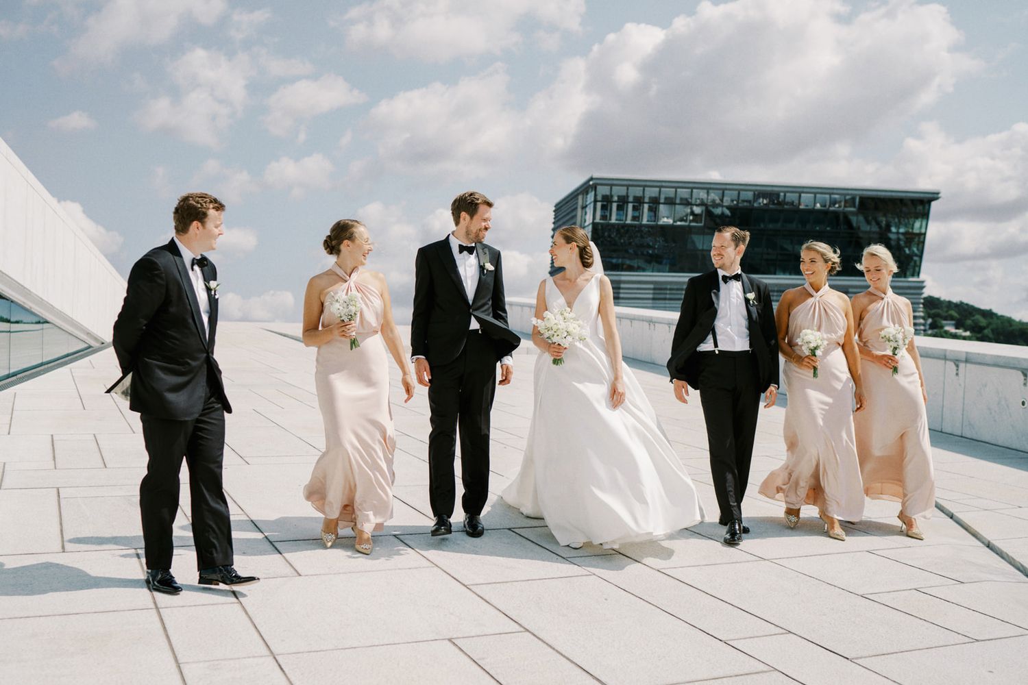 Wedding party walking on Oslo Opera House rooftop, bridesmaids in blush gowns and groomsmen in black tuxedos.