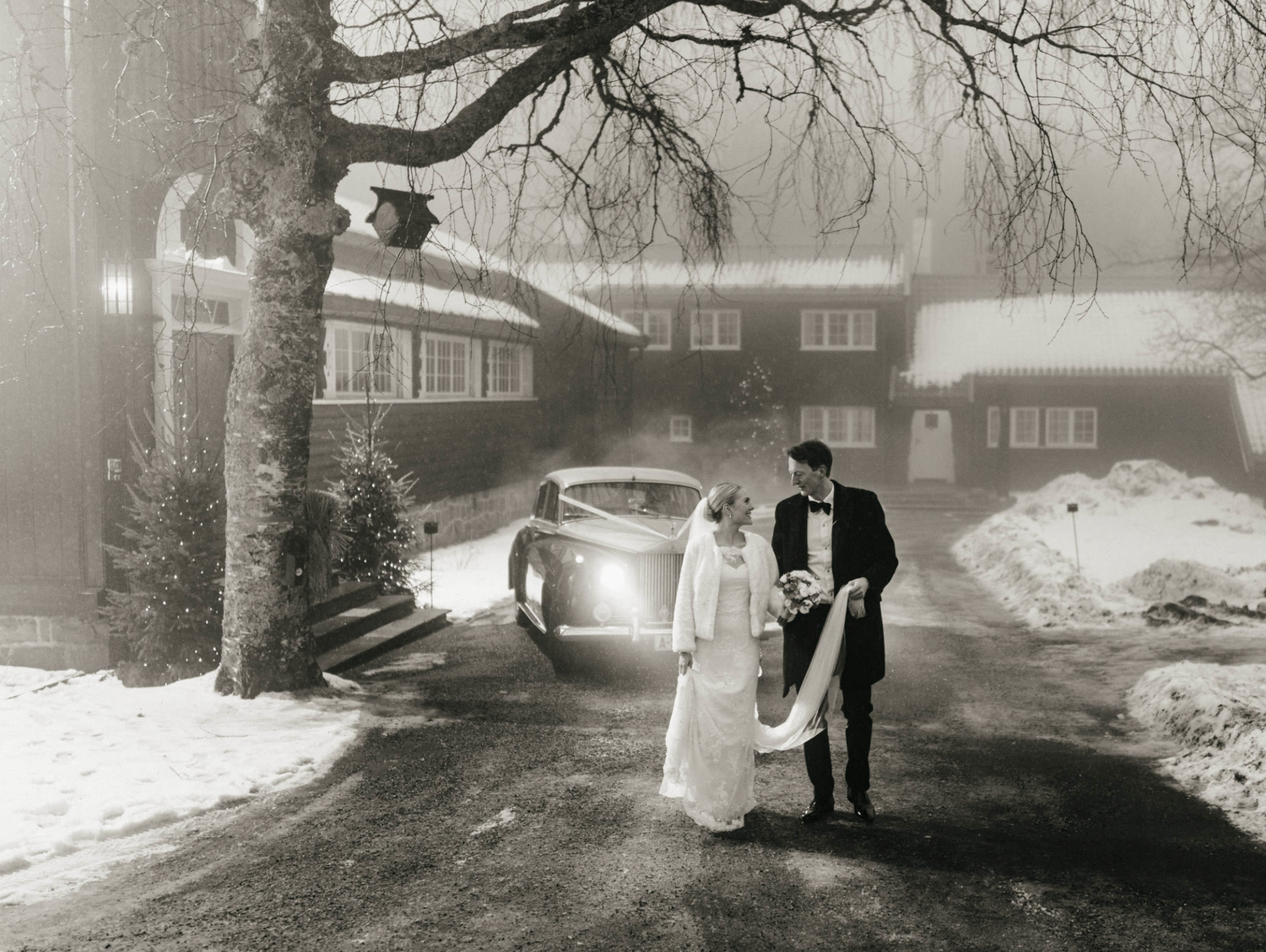Bride and groom walking in snowy night landscape beside vintage car in black and white photo.