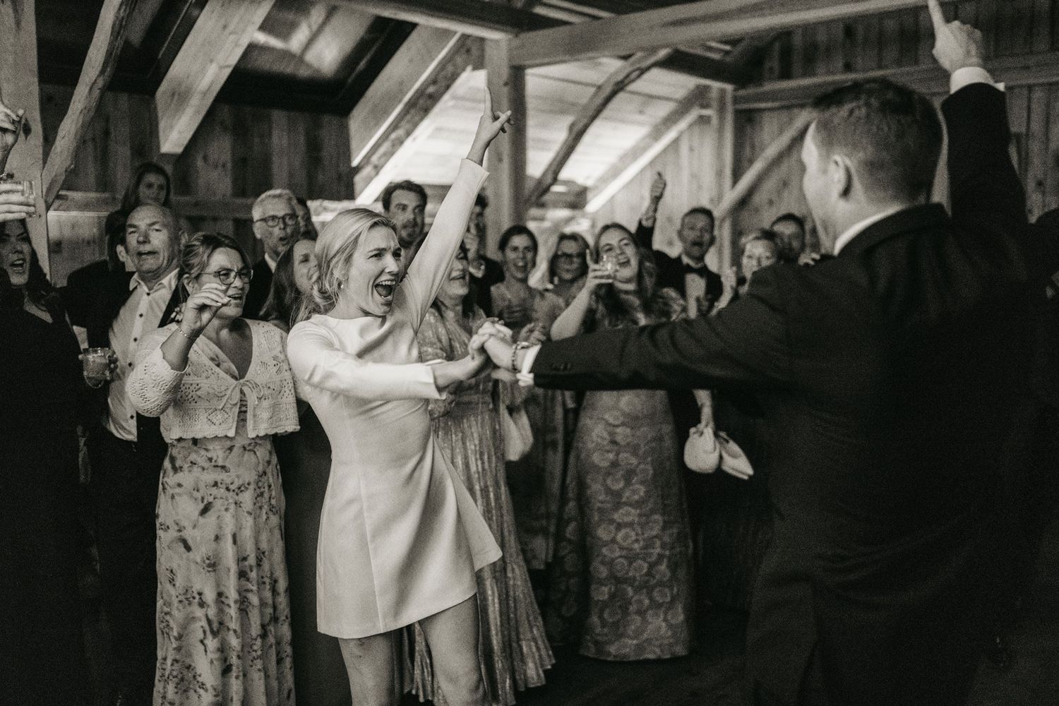 Bride and groom celebrate joyfully with guests clapping at rustic barn wedding reception in black and white photo.