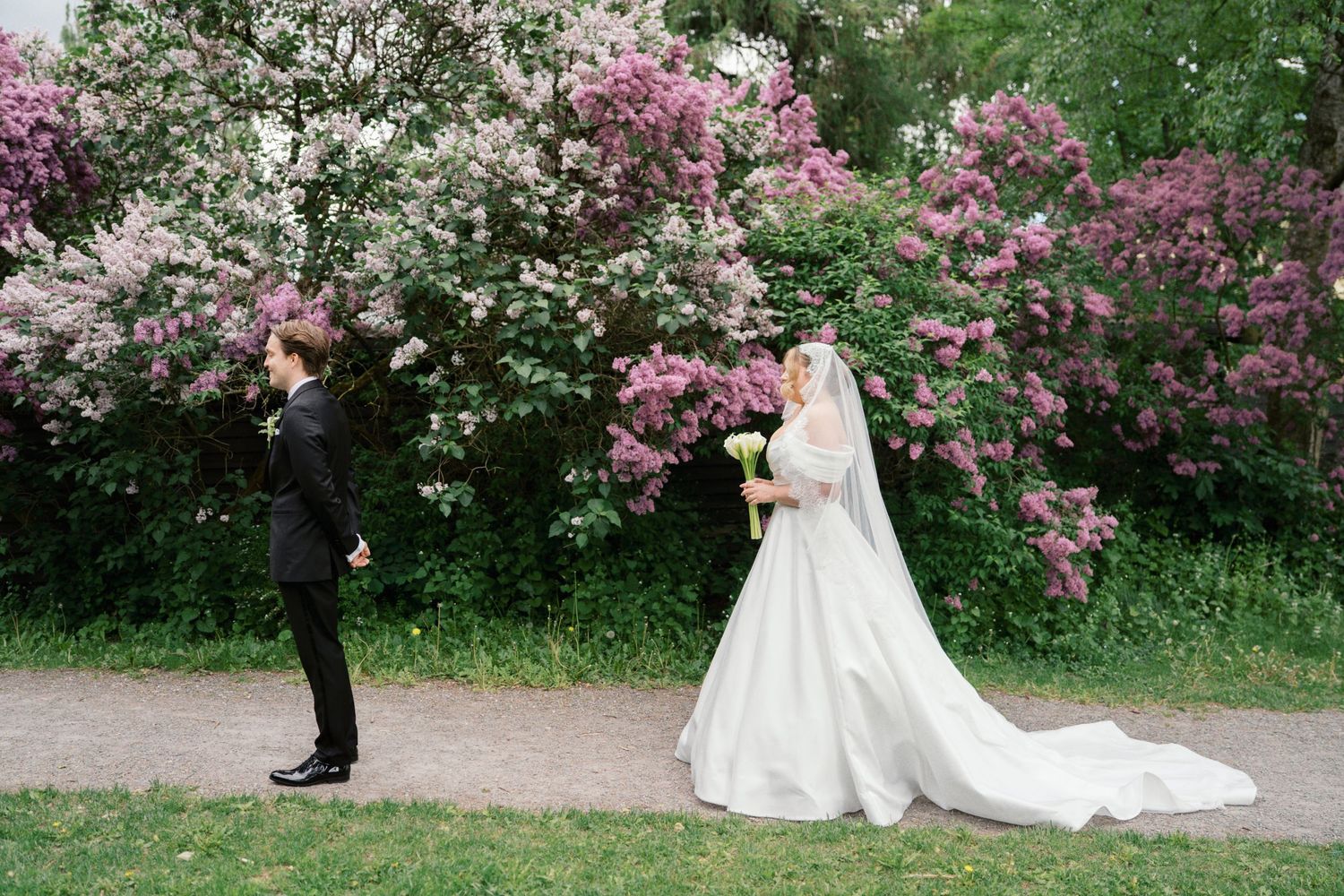 Bride in white gown and groom in black suit share first look near blooming pink lilac bushes in a garden.