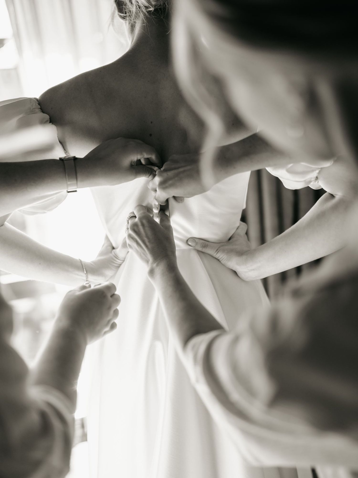 Black and white photo of bridesmaids helping a bride button up her white wedding dress before the ceremony.