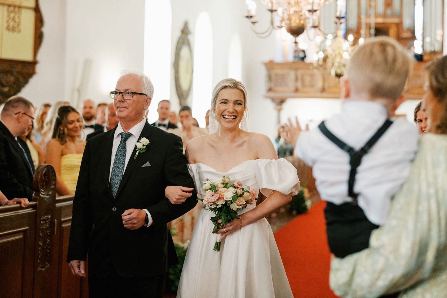 Bride walks down church aisle with older gentleman, holding pink bouquet, smiling at groom in elegant wedding ceremony.