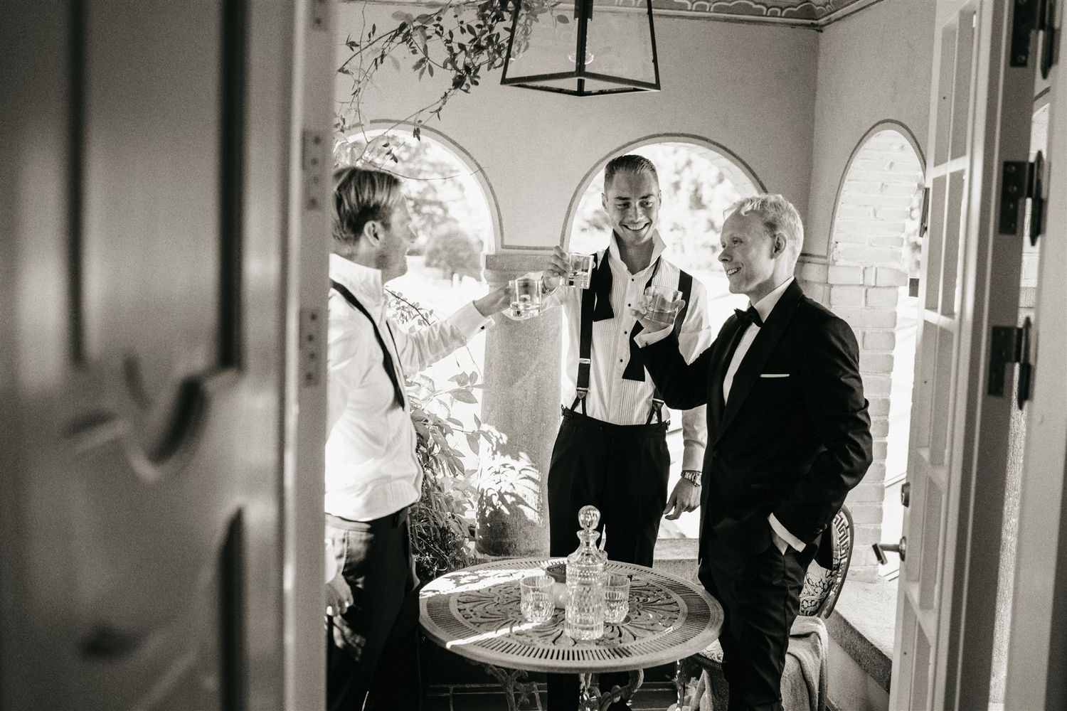 Black and white candid photo of groom and groomsmen sharing a toast and laughing in a sunlit doorway.