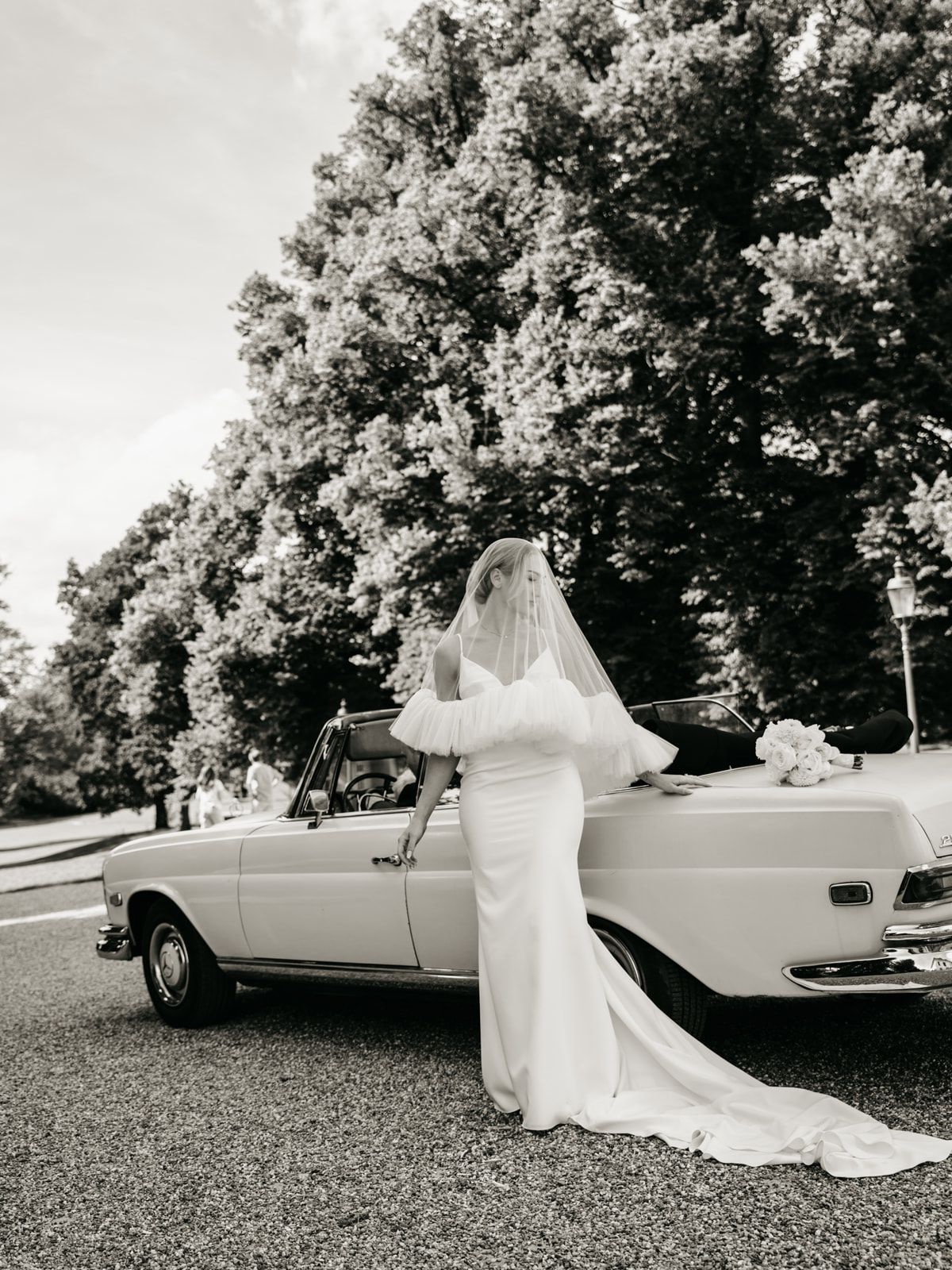 Elegant bride in fitted wedding gown and veil posing beside vintage convertible car, black and white.