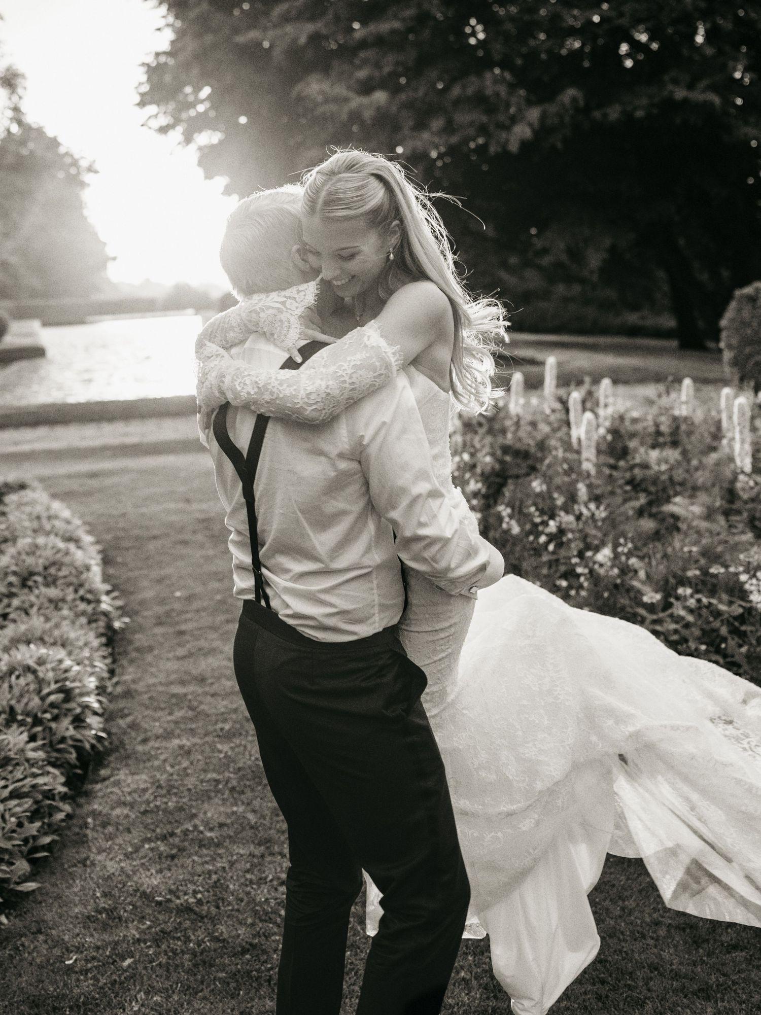 Black and white photo of a bride and groom embracing in a sunlit garden, the groom lifting his bride in a romantic moment.