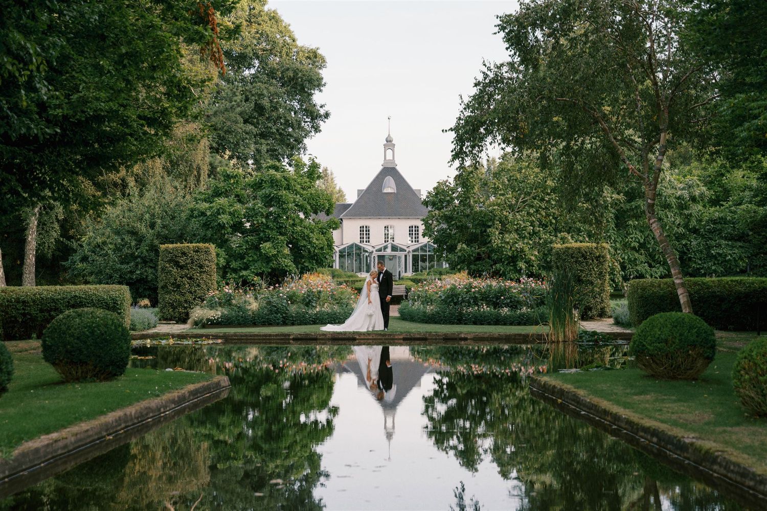Bride and groom reflected in garden pond before a charming French chateau estate.