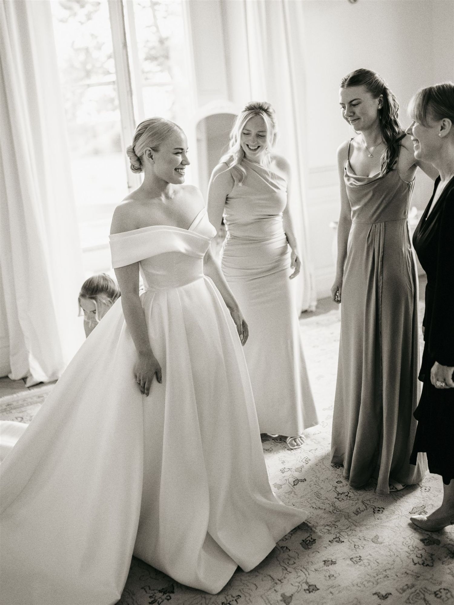 Black and white photo of bride in off-shoulder ballgown surrounded by smiling bridesmaids during bridal prep.