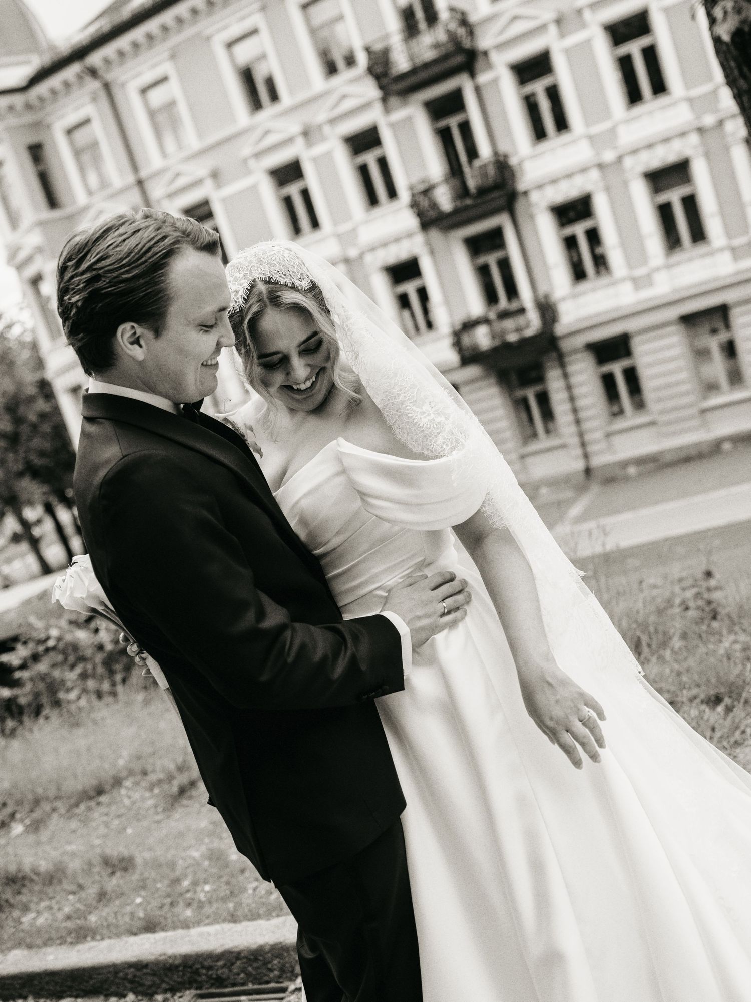 Black and white wedding photo of a smiling bride in white gown and groom in tuxedo embracing outdoors near a classic building.