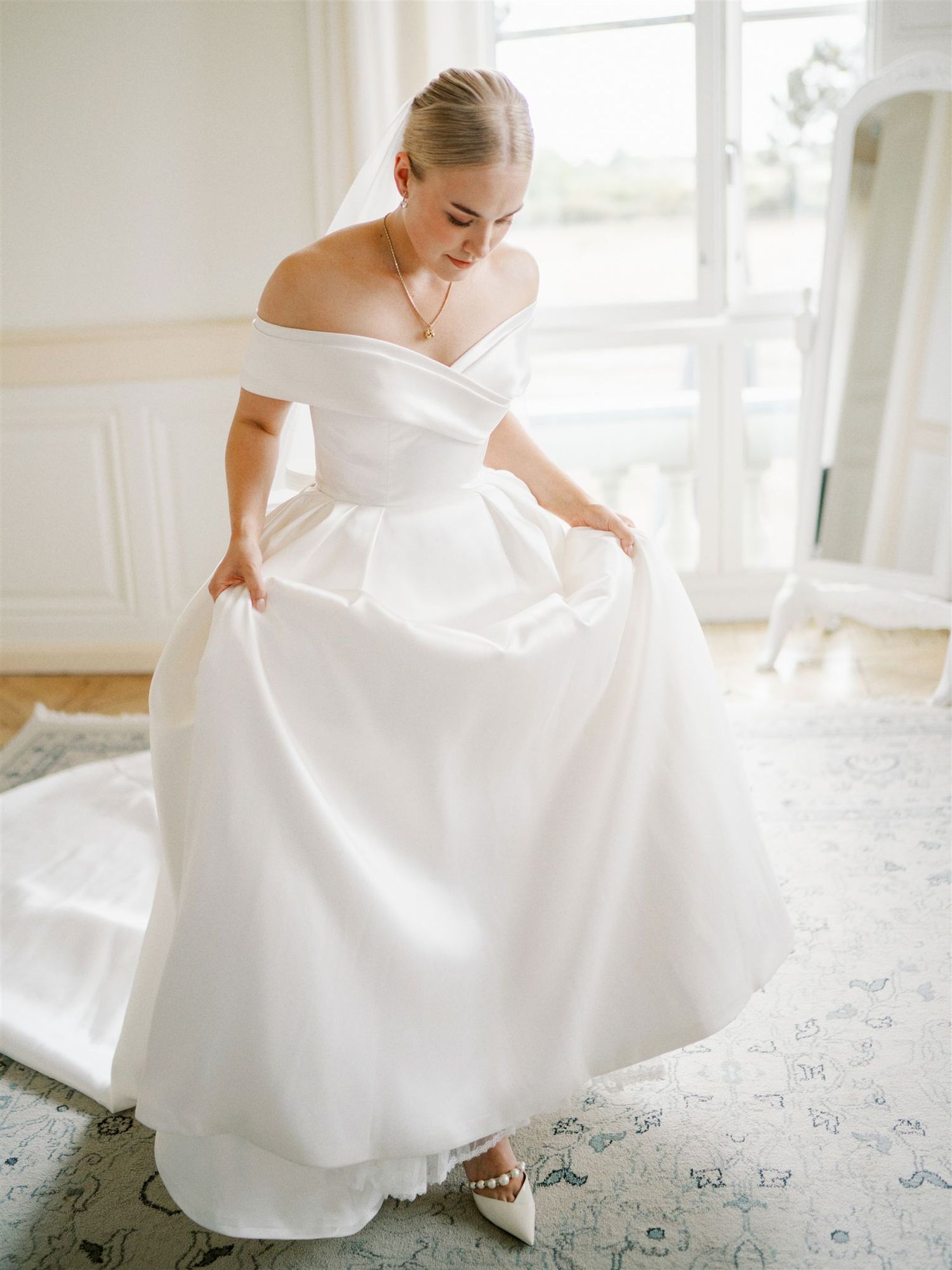 Bride in elegant off-shoulder white satin ball gown wedding dress adjusting her skirt indoors.