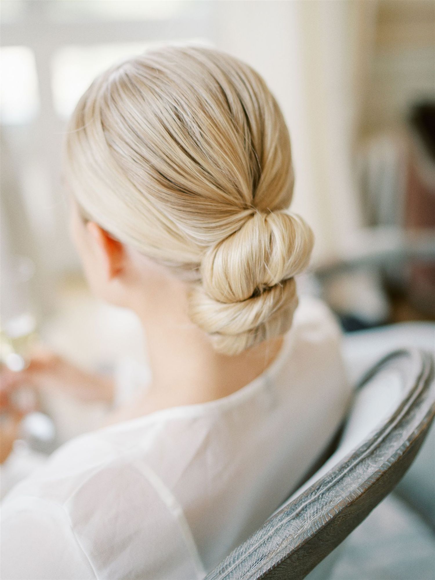 Elegant blonde bride with sleek low chignon updo hairstyle wearing white robe, soft natural light.