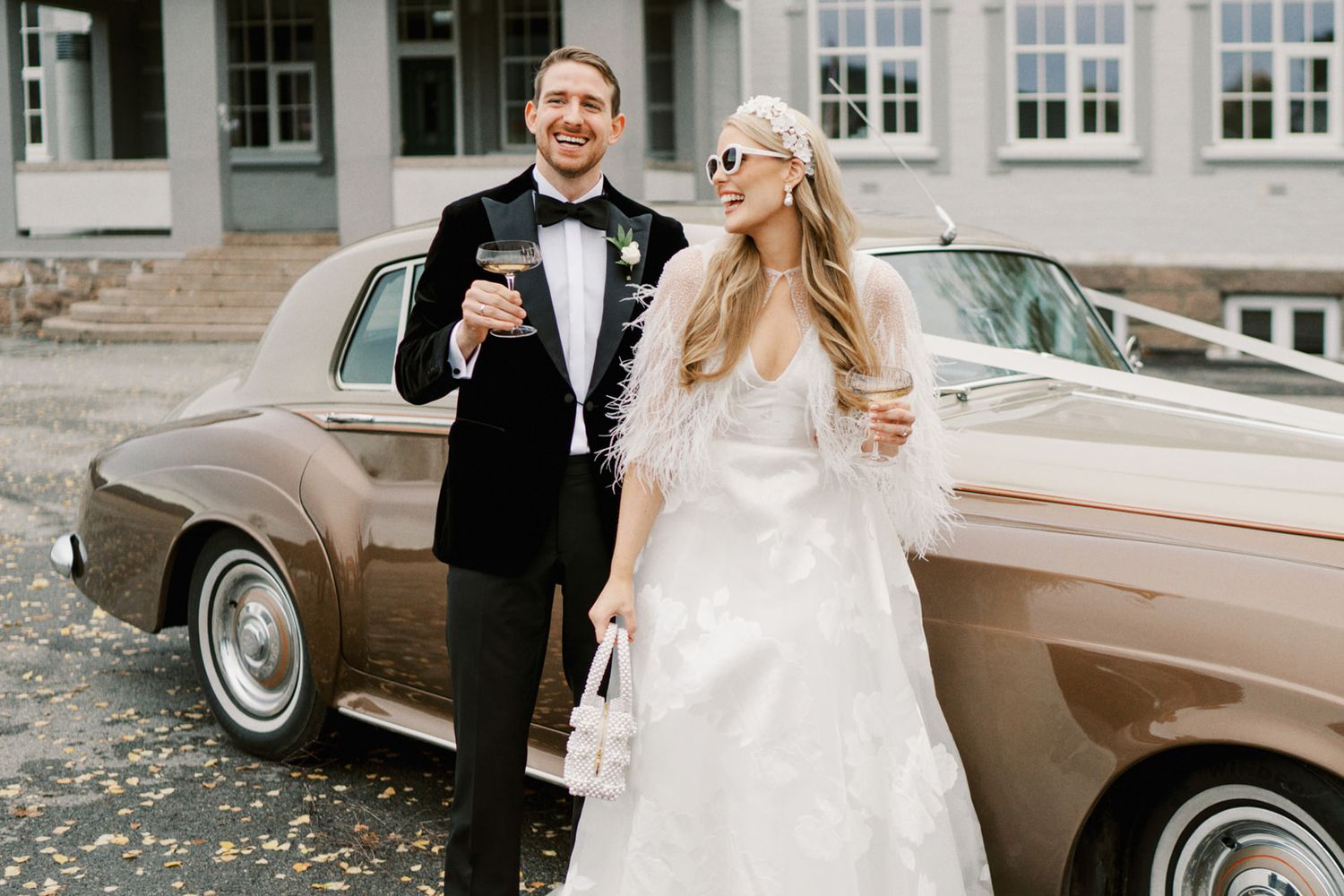 Smiling bride and groom holding champagne glasses beside a vintage tan Rolls-Royce on wedding day.