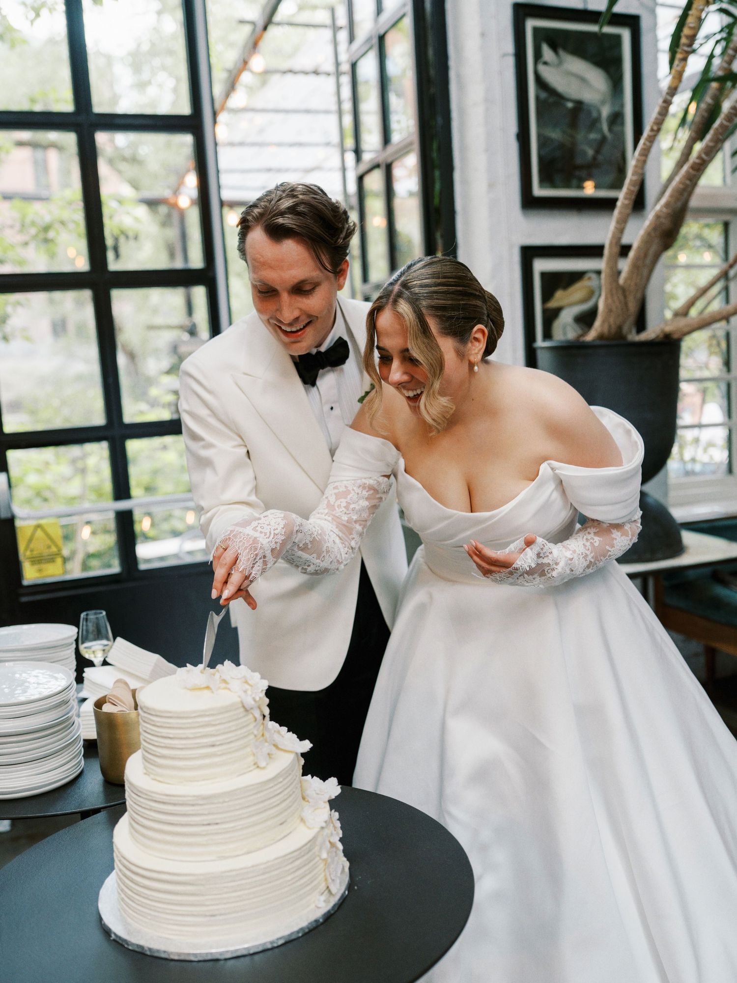 Bride and groom cutting white tiered wedding cake in elegant greenhouse venue, groom in white tuxedo.