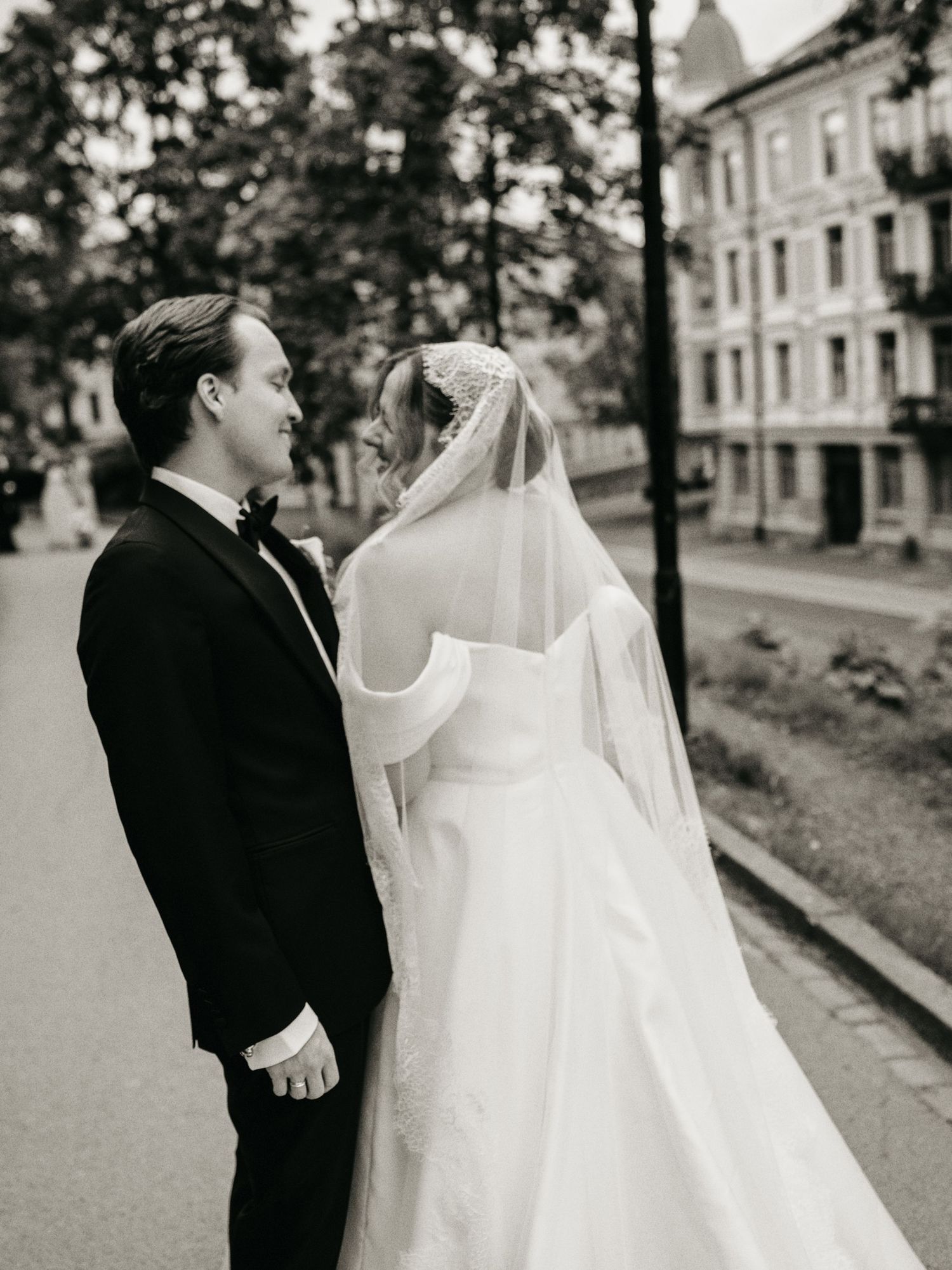 Black and white wedding photo of bride in off-shoulder gown with veil and groom in tuxedo standing on a tree-lined street.