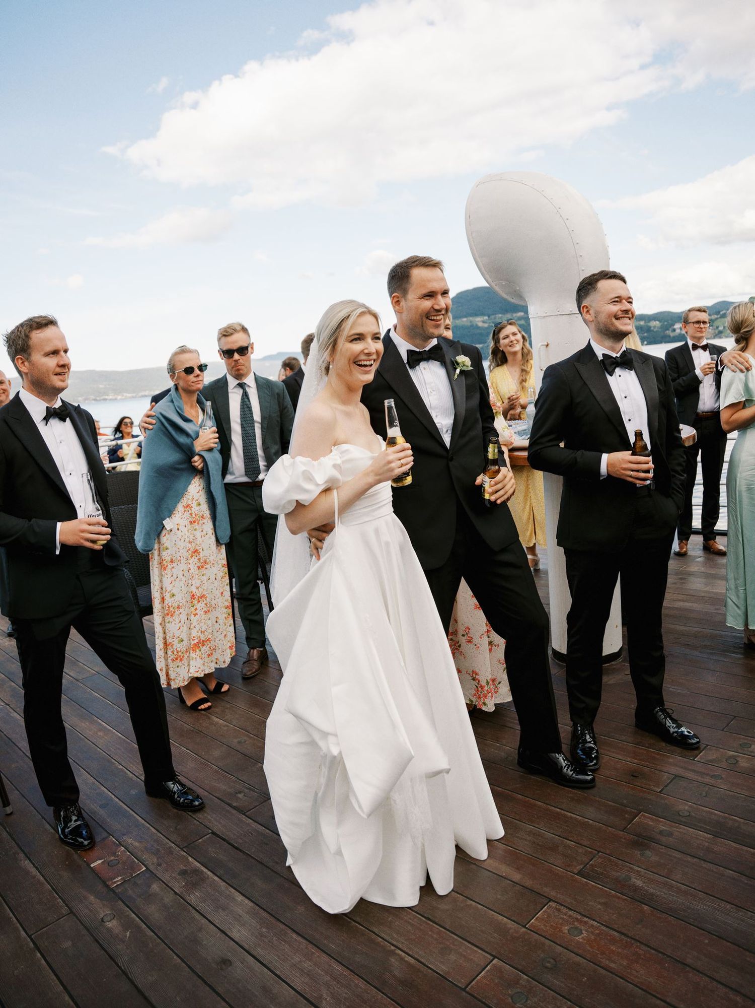 Bride in white off-shoulder gown and groom in tuxedo celebrate with guests on ship deck holding champagne.