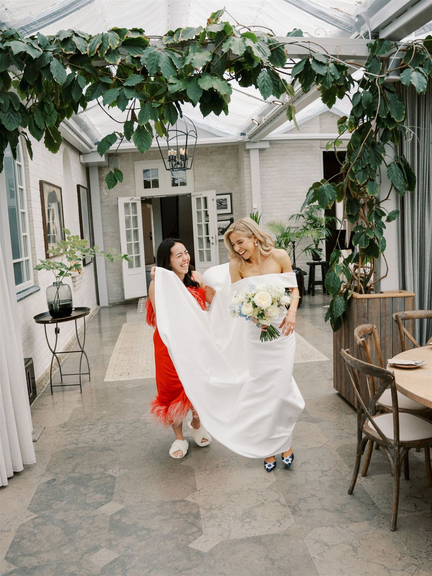 Bridesmaid in red dress helping bride carry white gown train in greenhouse venue.