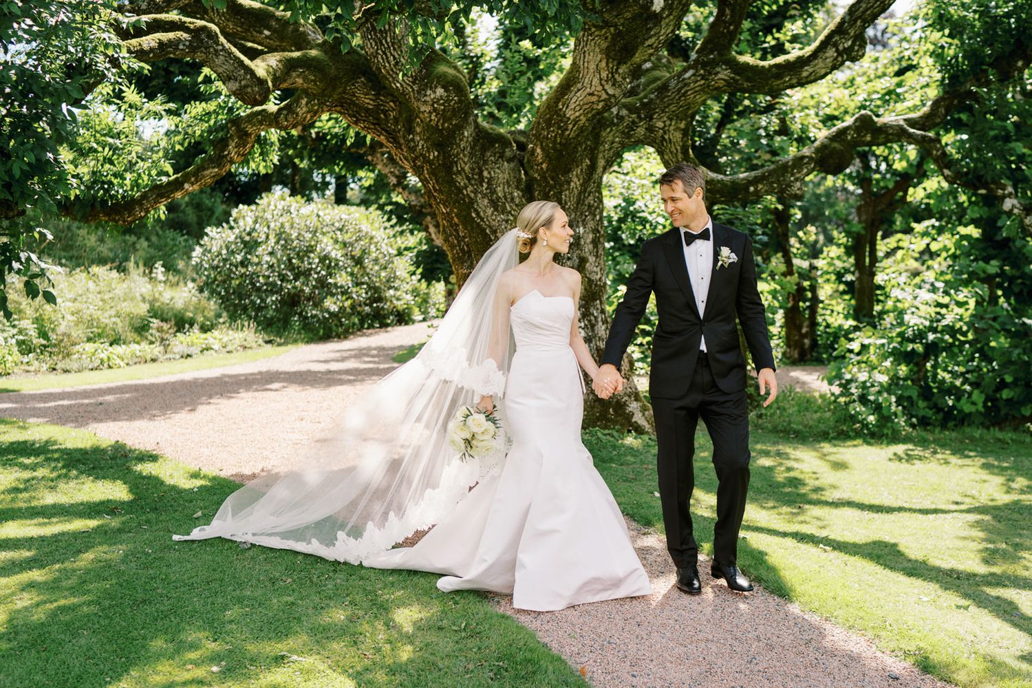 Bride in white gown with veil and groom in black tuxedo stroll hand in hand under large tree.