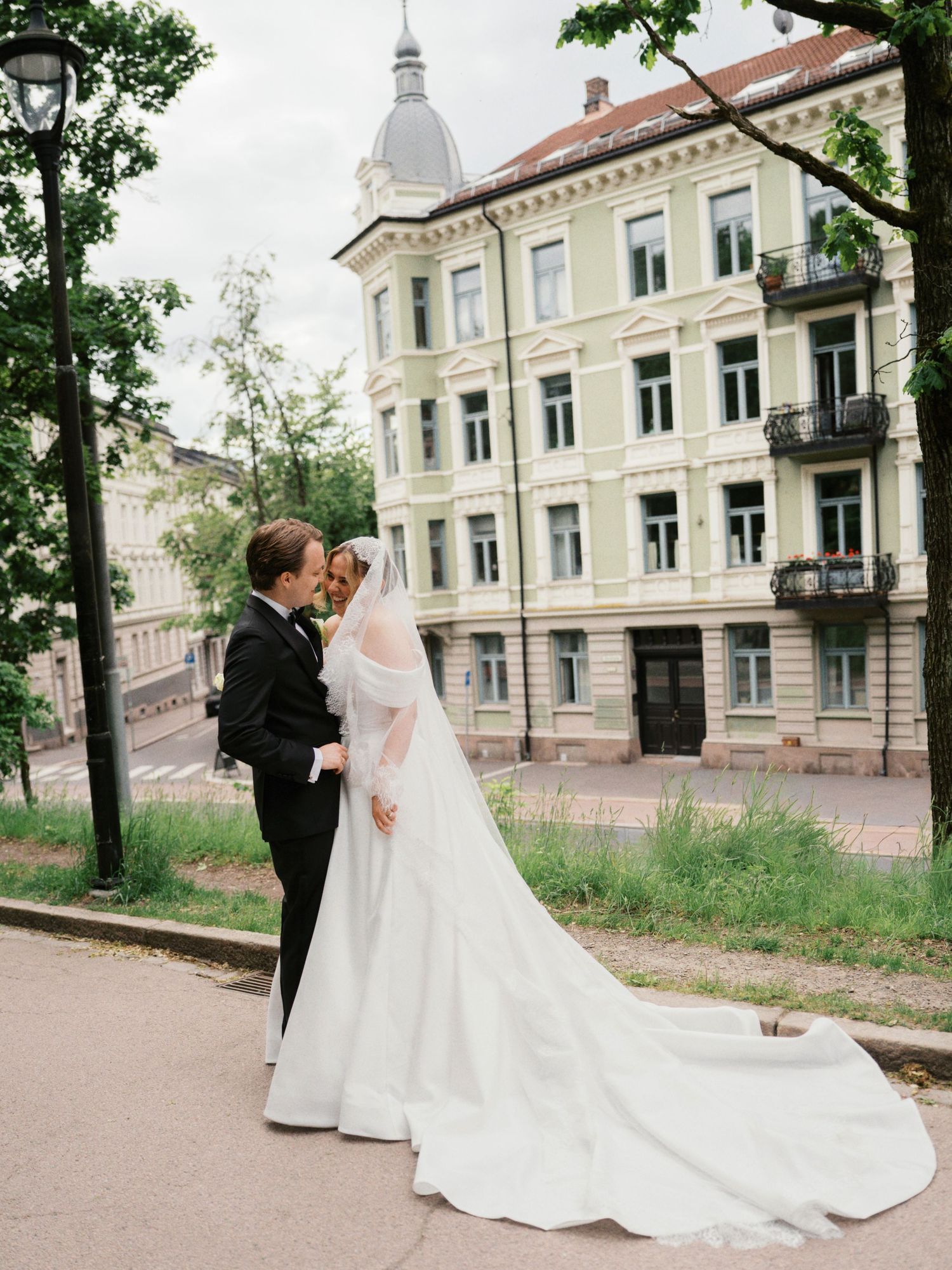 Bride in white gown with long veil and groom in black tuxedo pose on a city street with elegant European building.