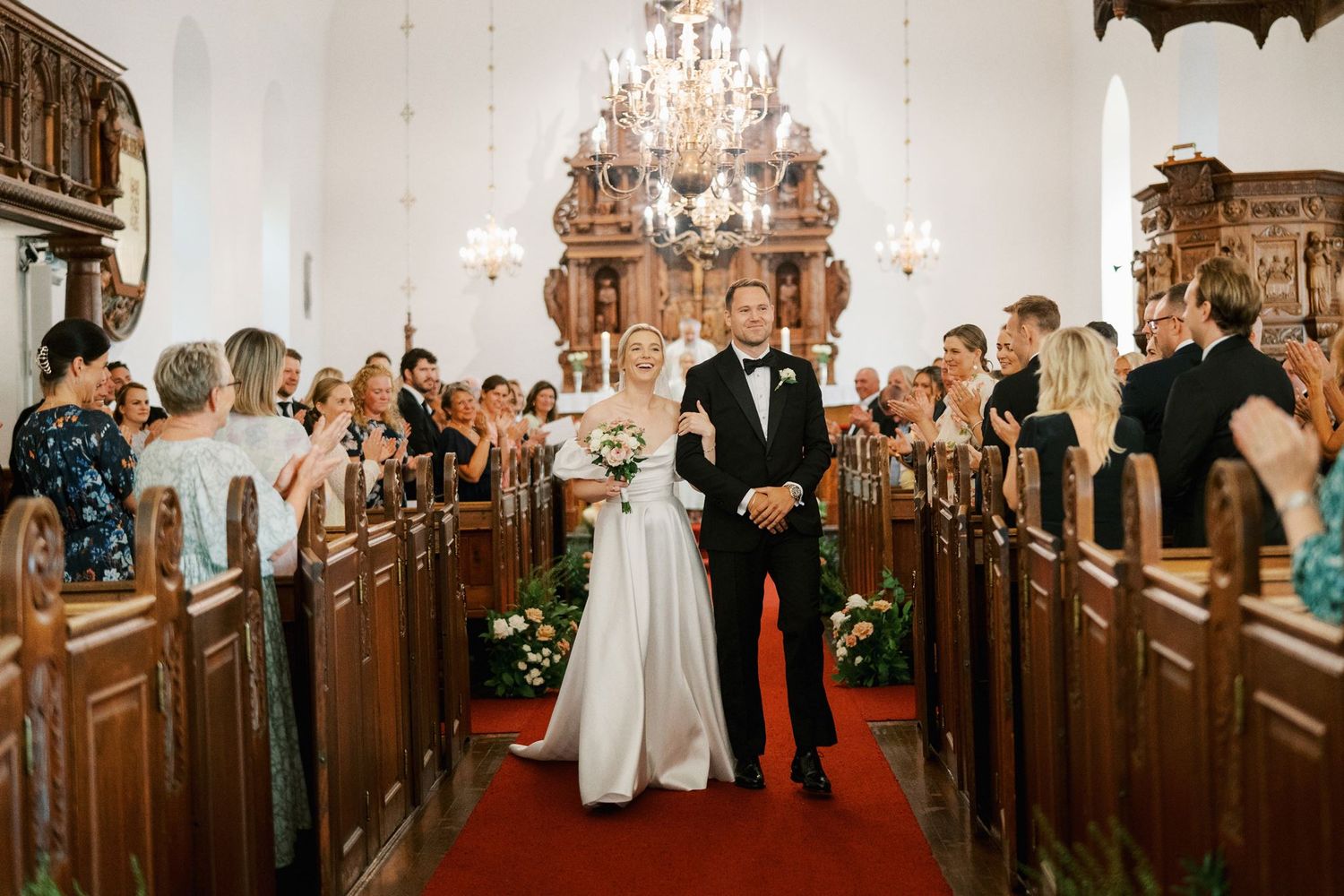 Bride and groom walk down red carpet aisle in elegant church wedding ceremony surrounded by guests.