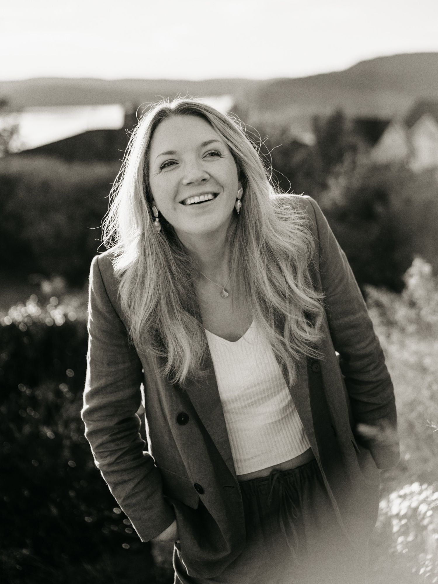 Black and white portrait of smiling blonde woman in blazer outdoors with scenic hills in background.