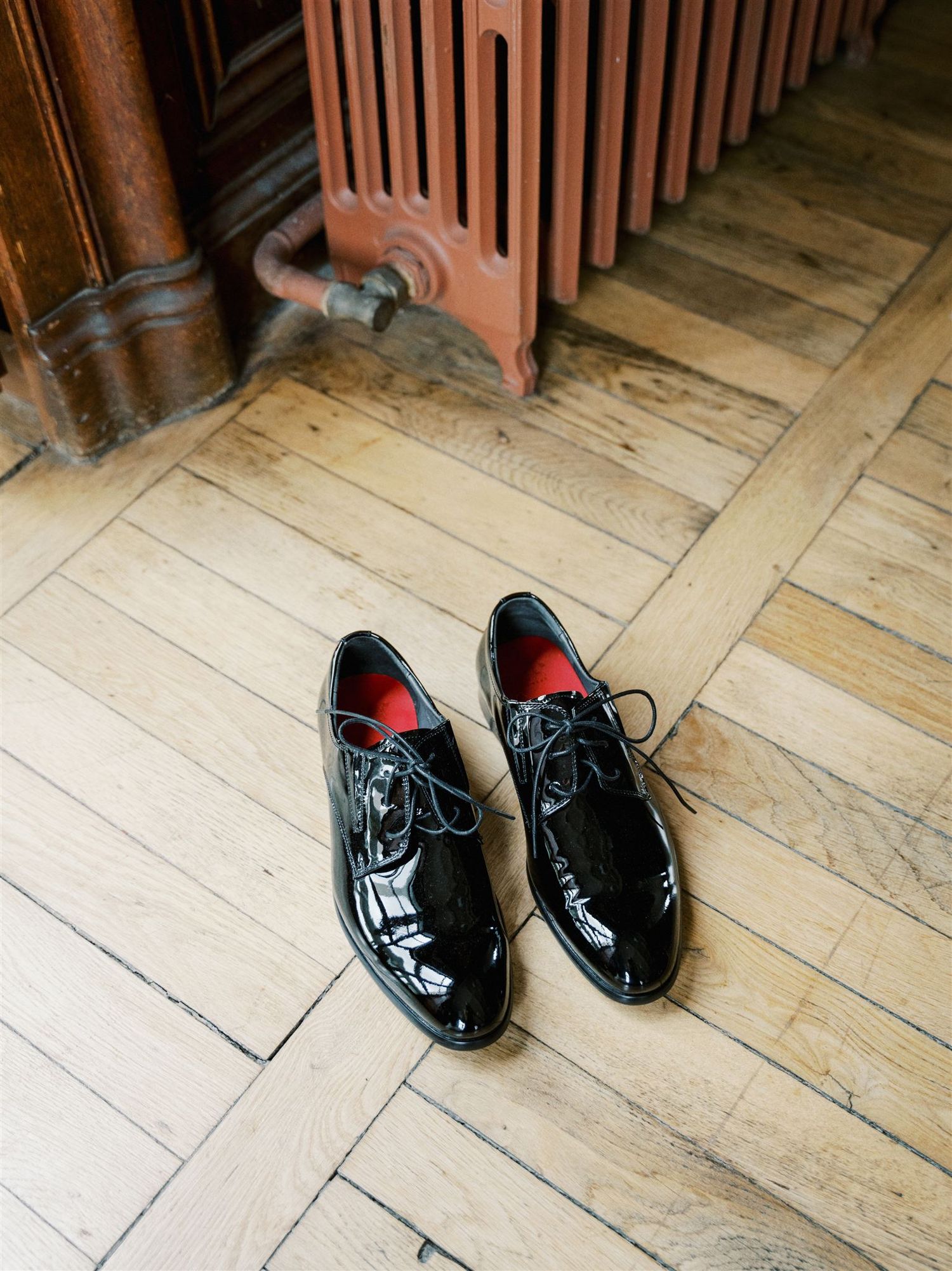 Shiny black patent leather Oxford shoes with red insoles placed on herringbone parquet wooden floor.