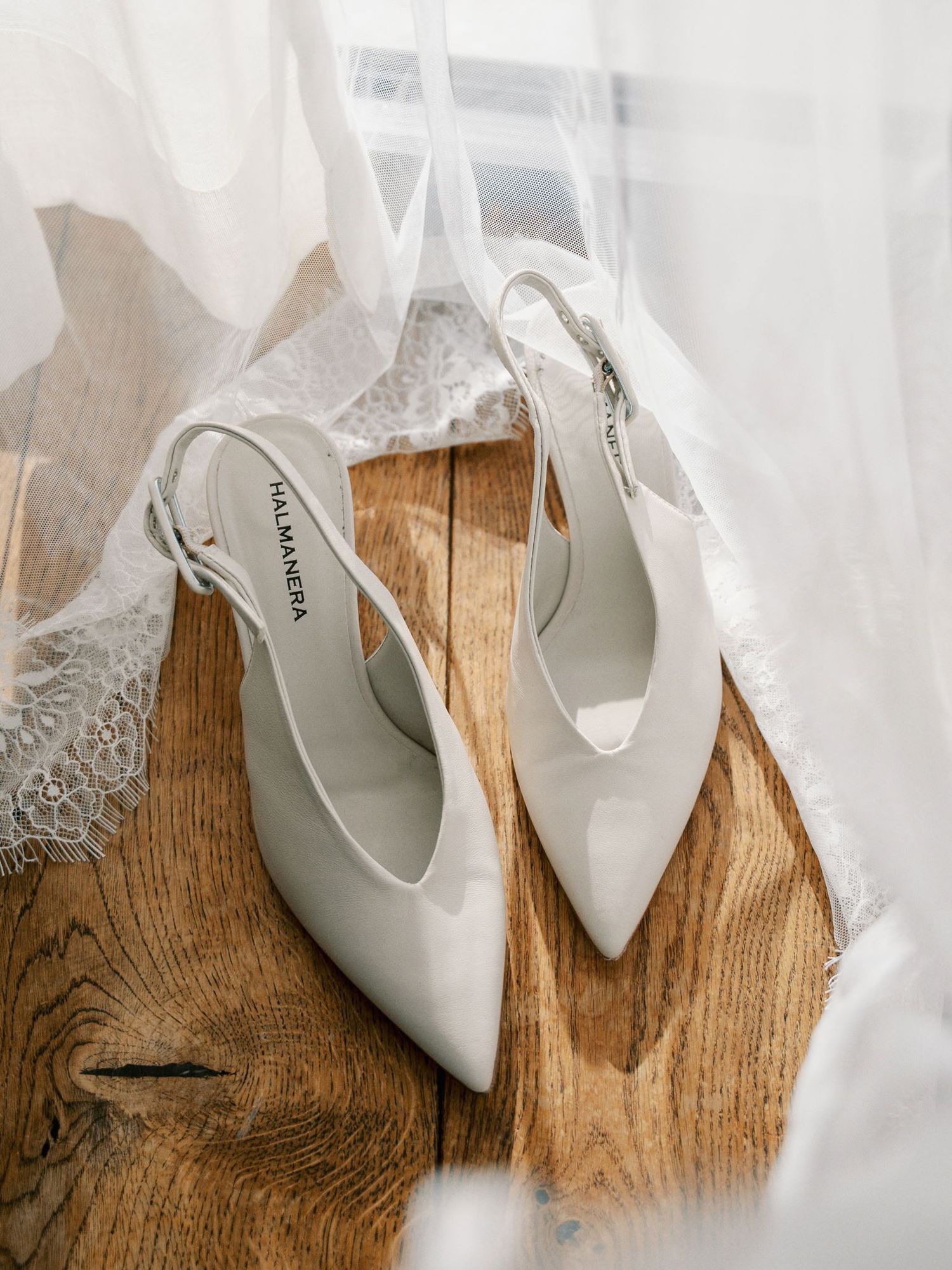 Elegant white slingback heels by Halmanera placed on rustic wood floor with bridal veil draped around them.
