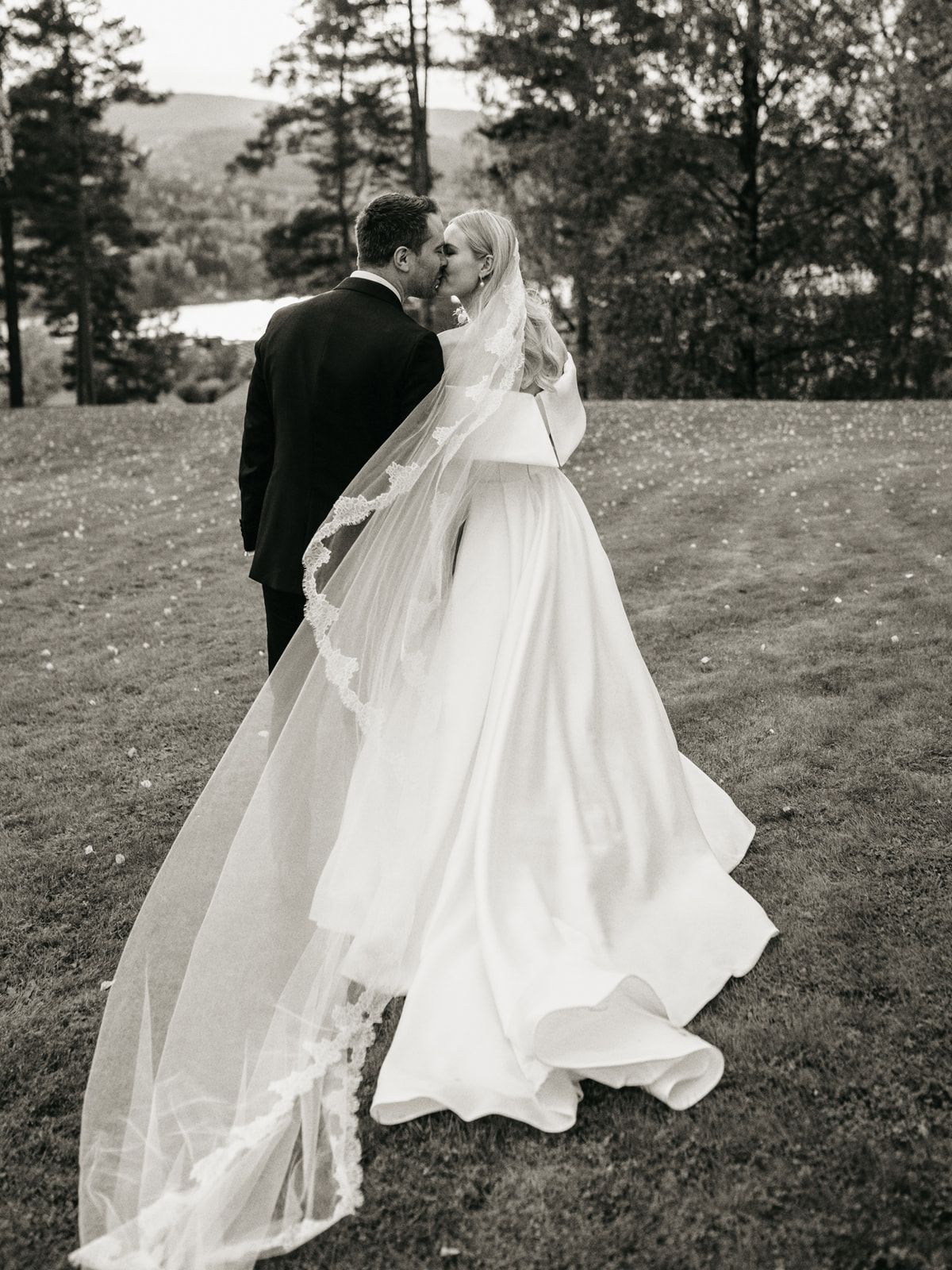 Black and white photo of bride in flowing gown and veil with groom in tuxedo outdoors.