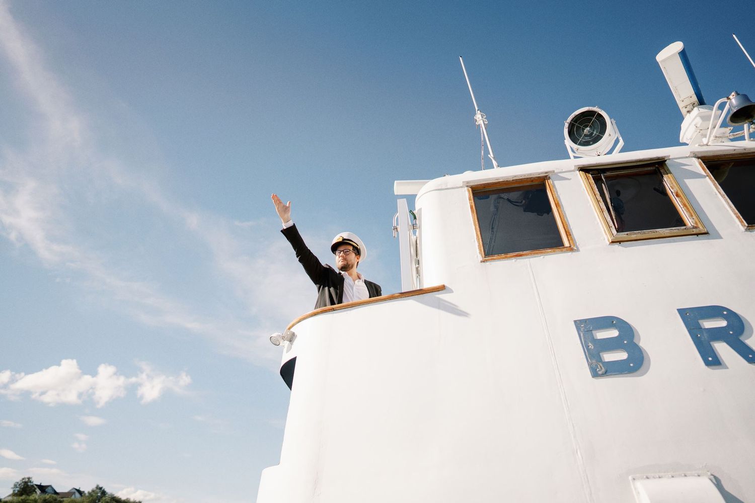 Person waving from the deck of a white boat named BR under a bright blue sky.