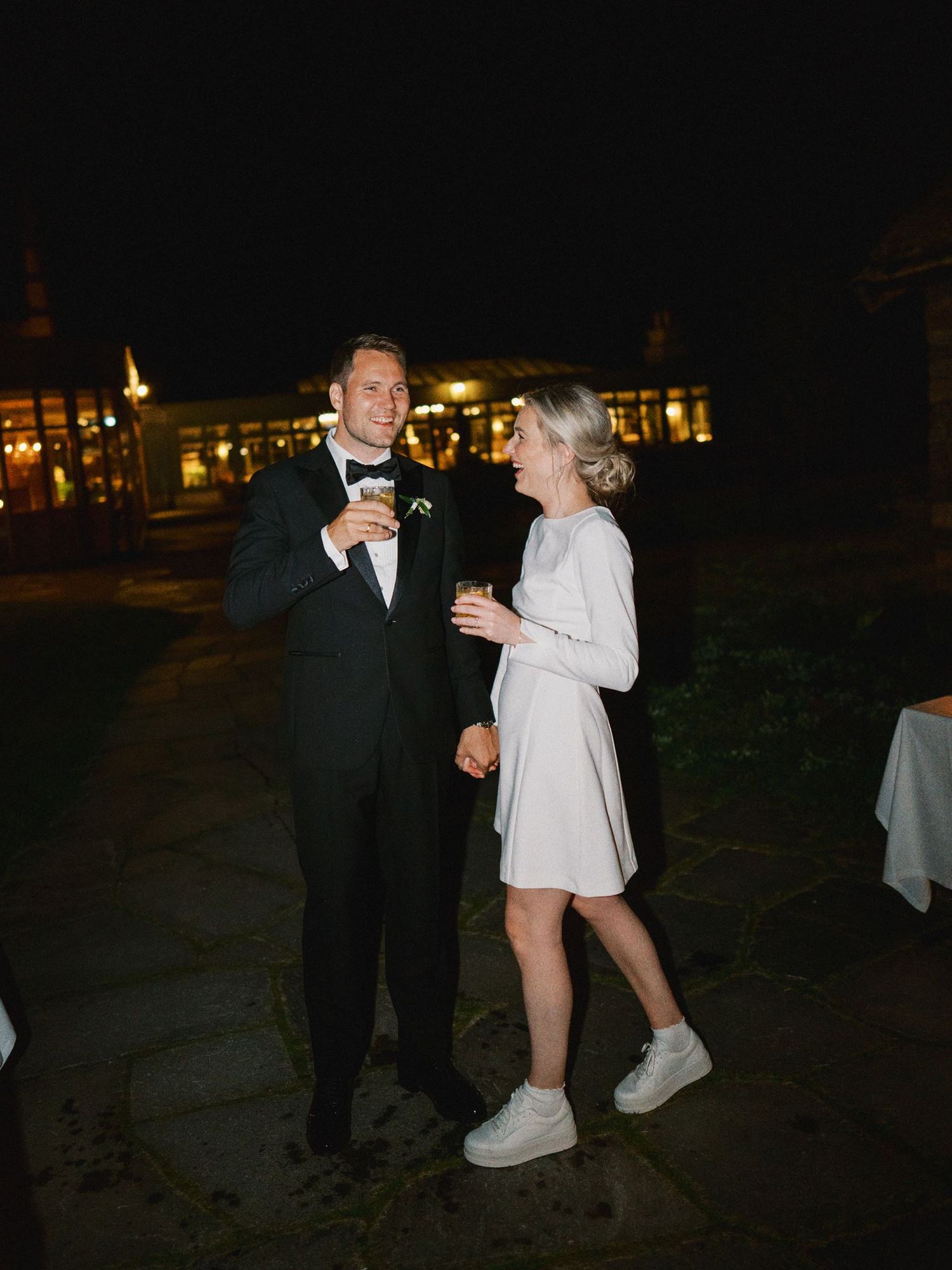 Couple dancing outdoors at night, man in tuxedo and woman in white dress with sneakers, elegant venue backdrop.