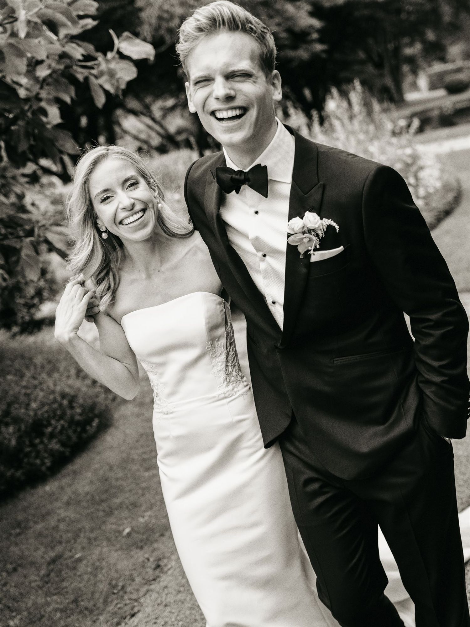 Black and white photo of smiling bride and groom in tuxedo at outdoor wedding.