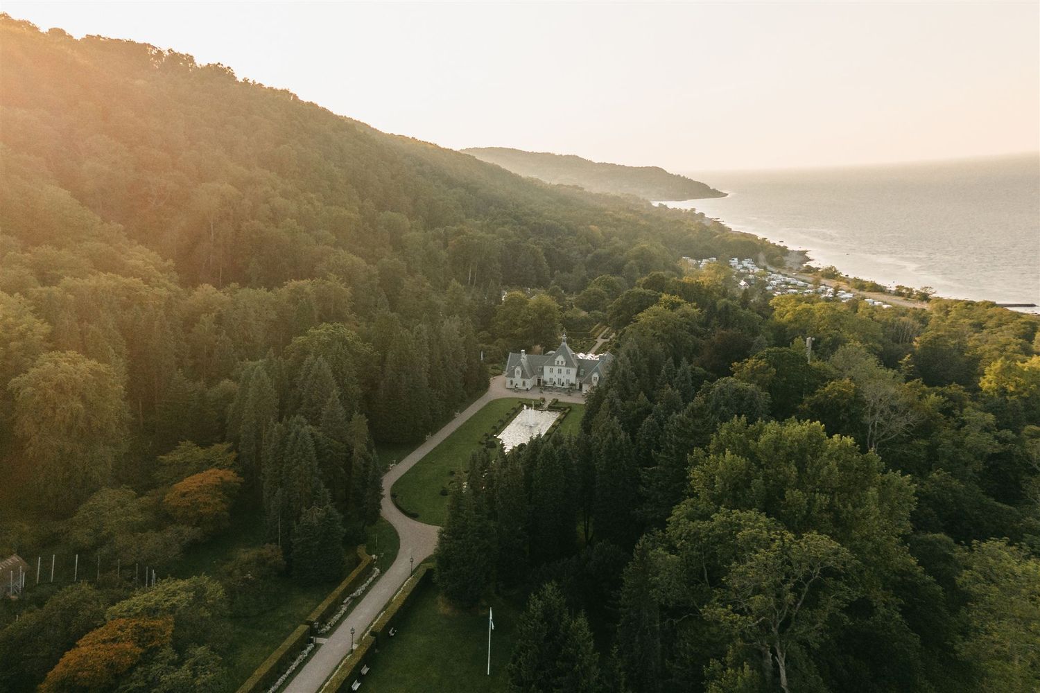 Aerial drone view of a grand estate nestled among lush green trees beside a shimmering coastline at golden hour.