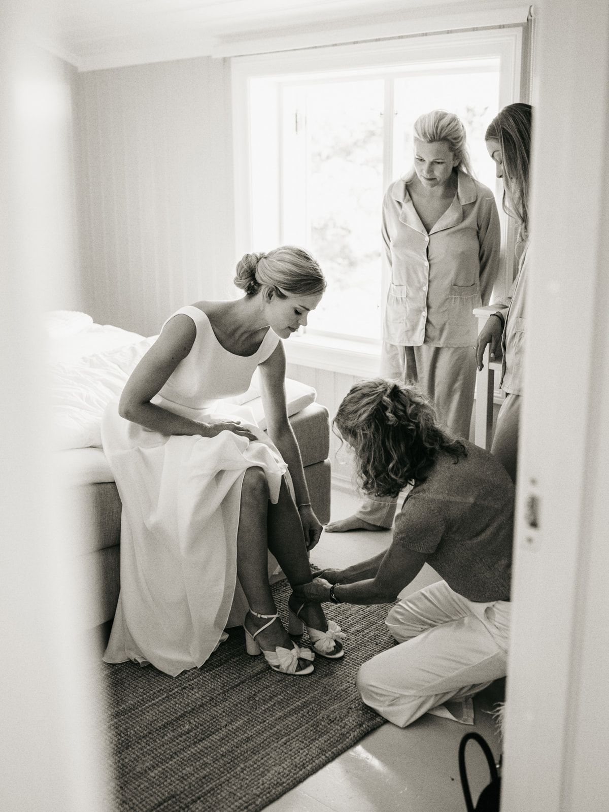 Bride in white gown getting shoes fastened by assistant while two women watch in bright bedroom.
