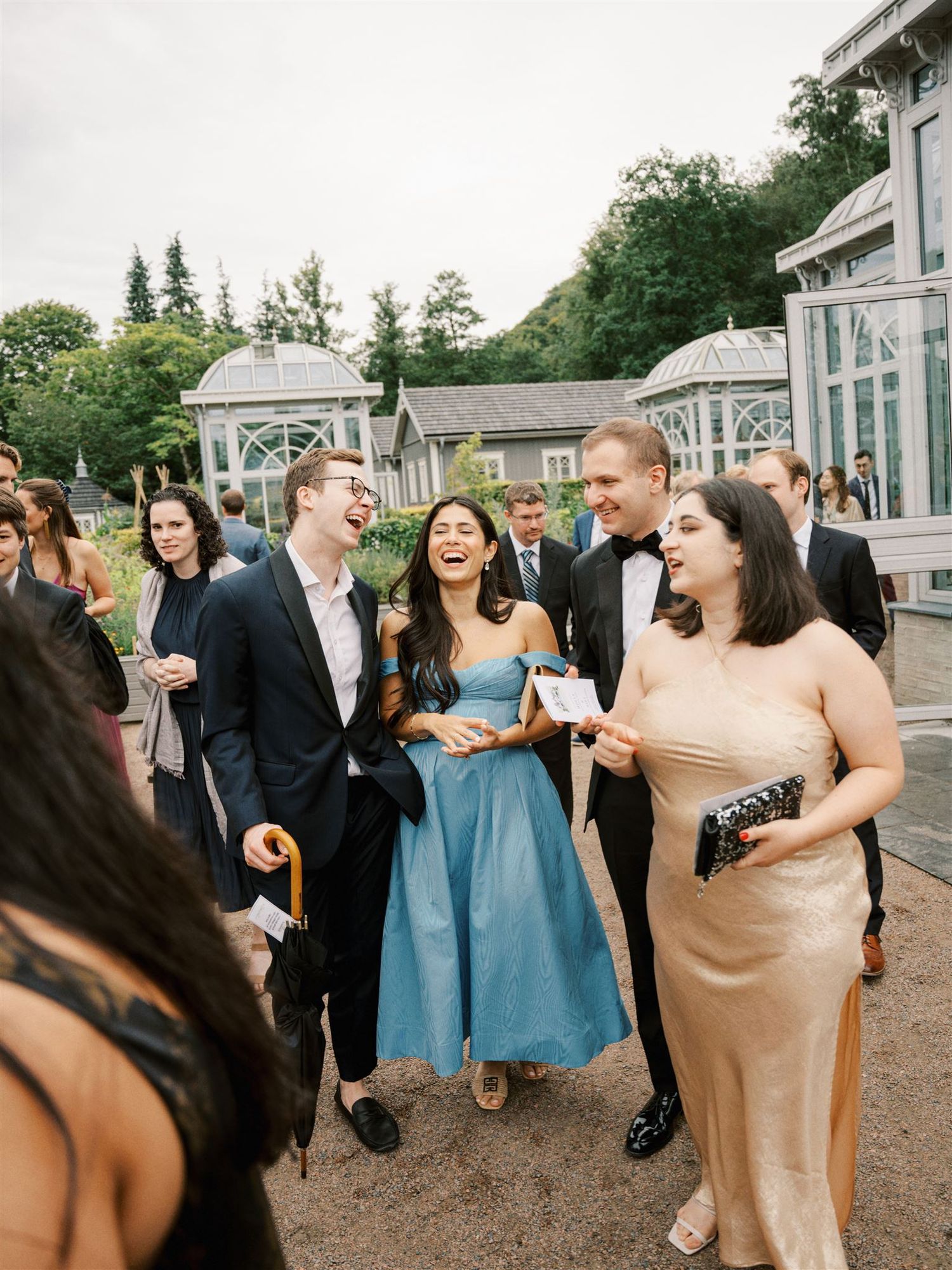 Wedding guests laughing and walking outdoors near elegant glass greenhouse venue.