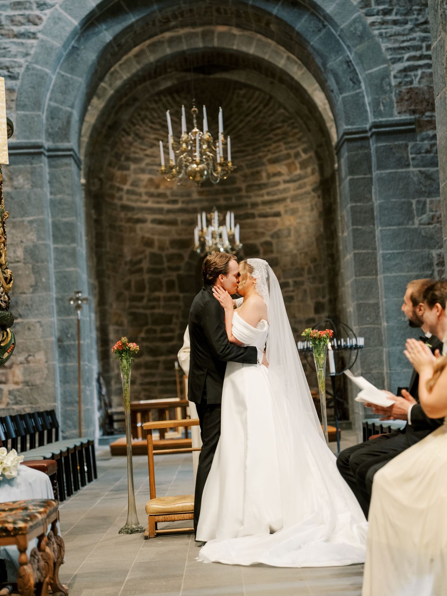 Bride and groom share first kiss at stone church wedding ceremony with chandelier and guests applauding.