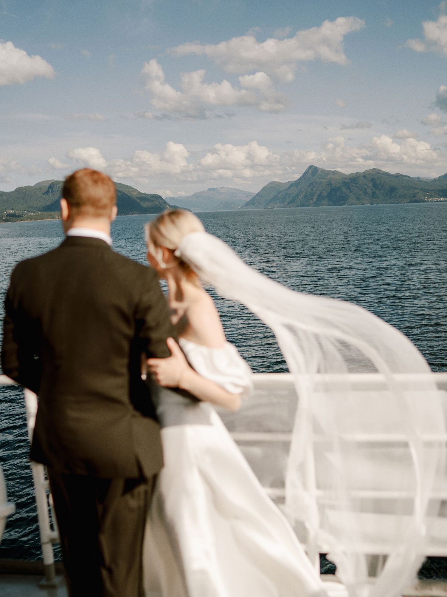 Bride and groom stand on a boat deck, her veil blowing in the wind, with stunning mountain lake views behind them.