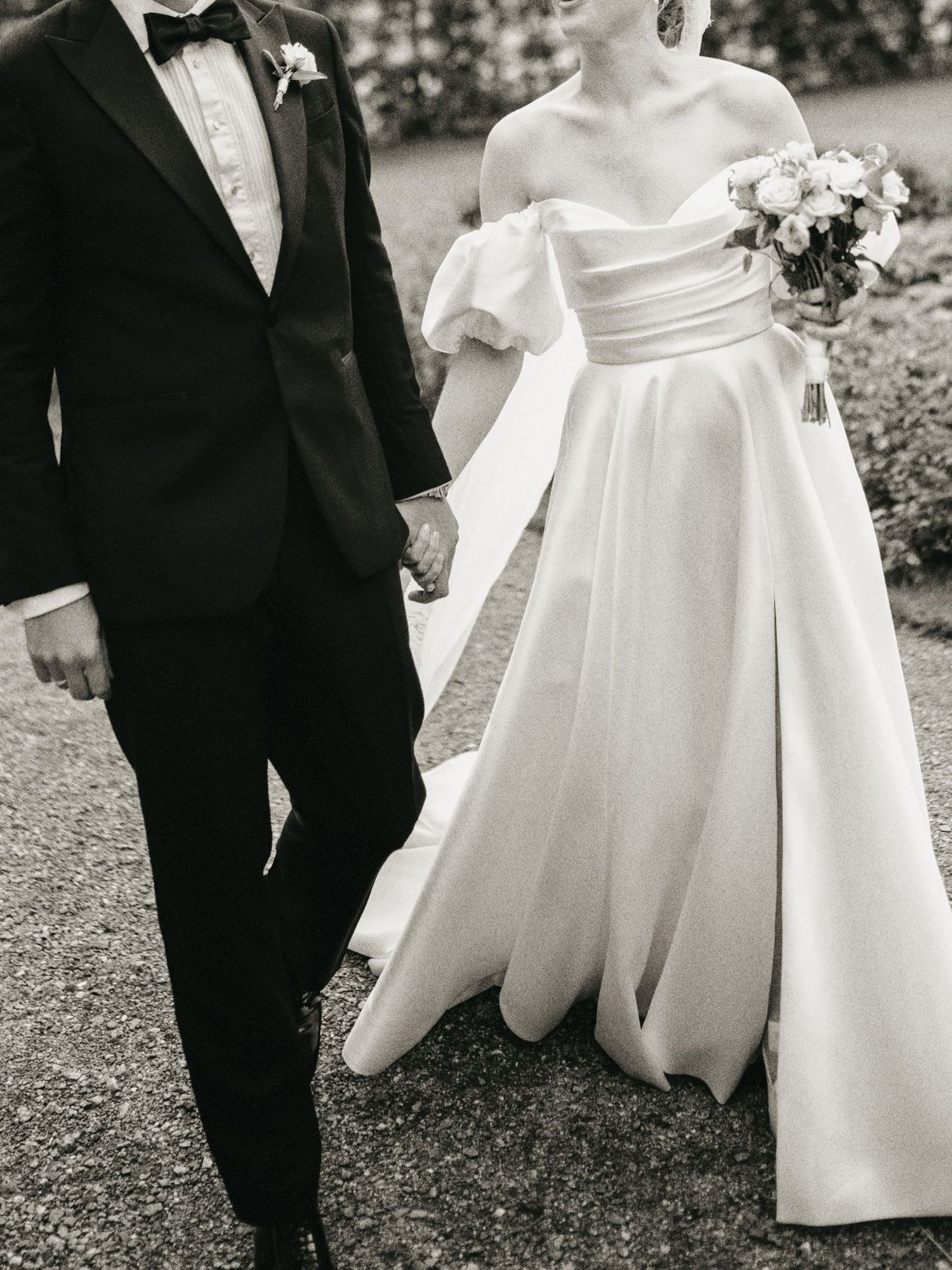 Black and white photo of bride in off-shoulder ball gown holding bouquet walking with groom in tuxedo.