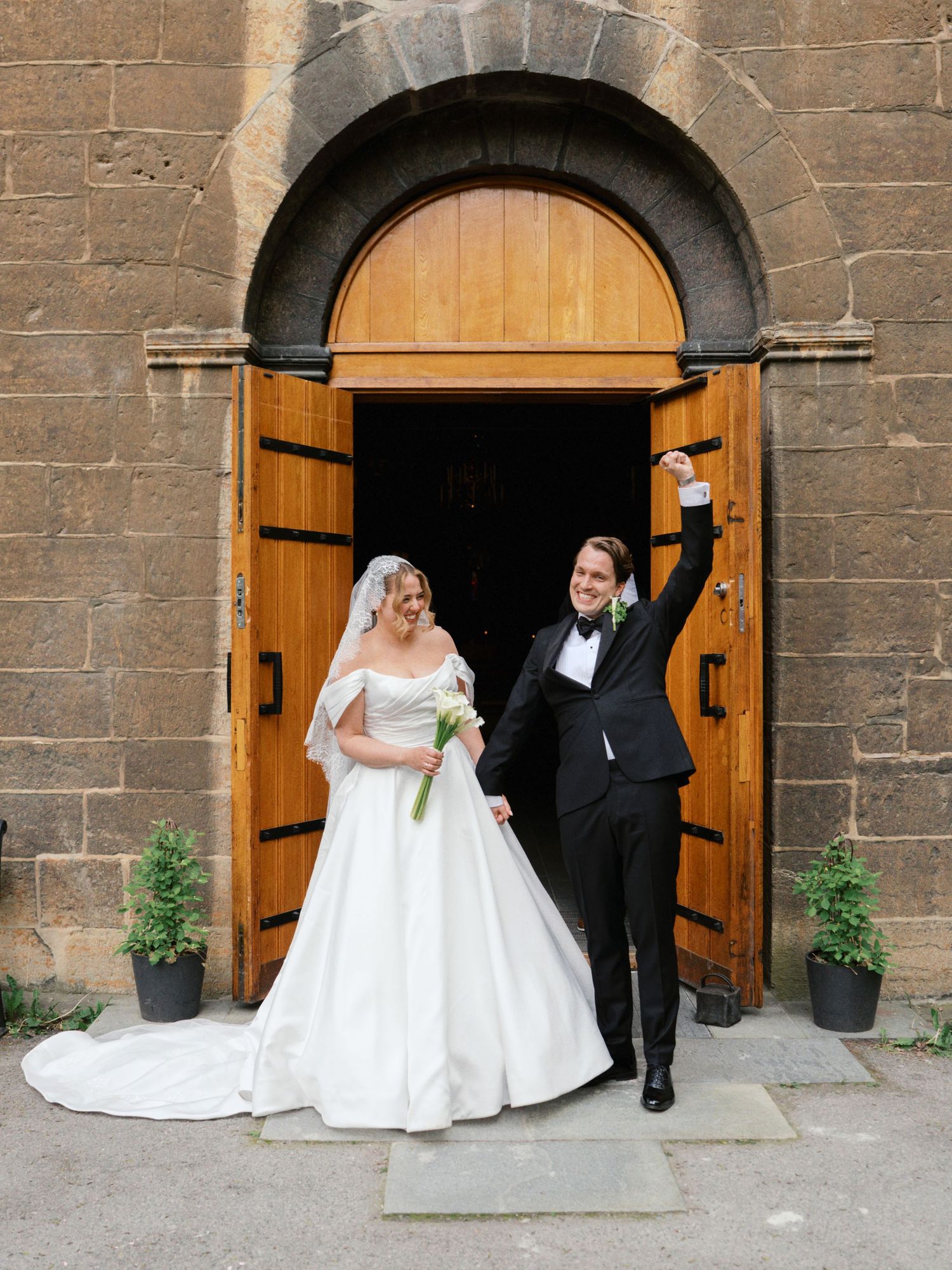 Bride and groom celebrating at church doors, bride in ball gown with veil, groom in tuxedo raising fist in joy.