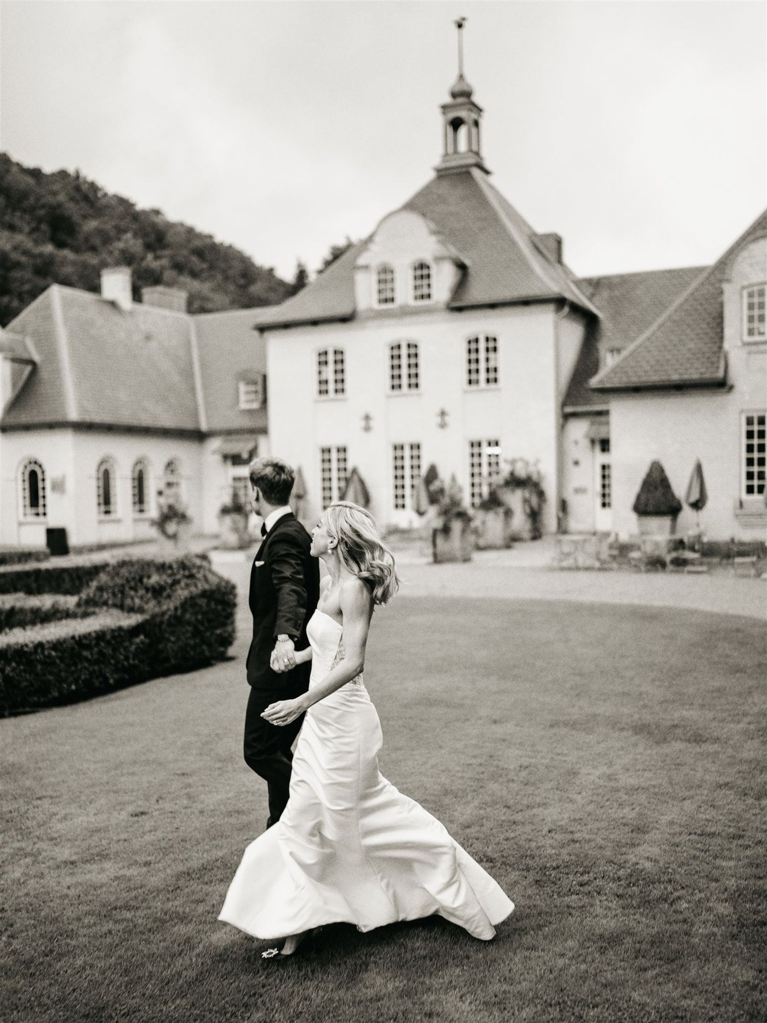 Black and white photo of bride in mermaid gown and groom walking hand in hand before an elegant manor house.