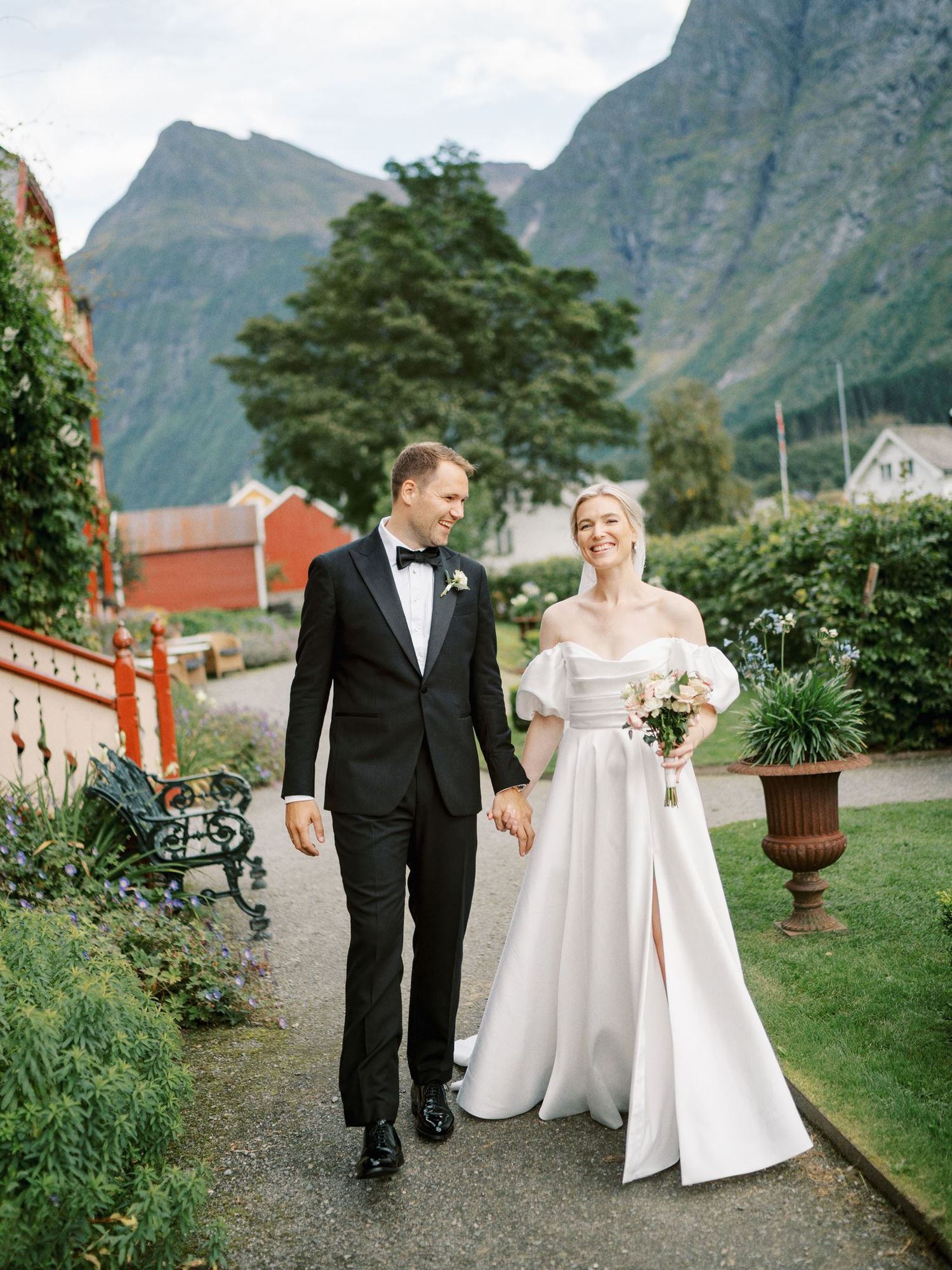 Bride and groom walk hand in hand through a lush garden with dramatic Norwegian mountains in the background.