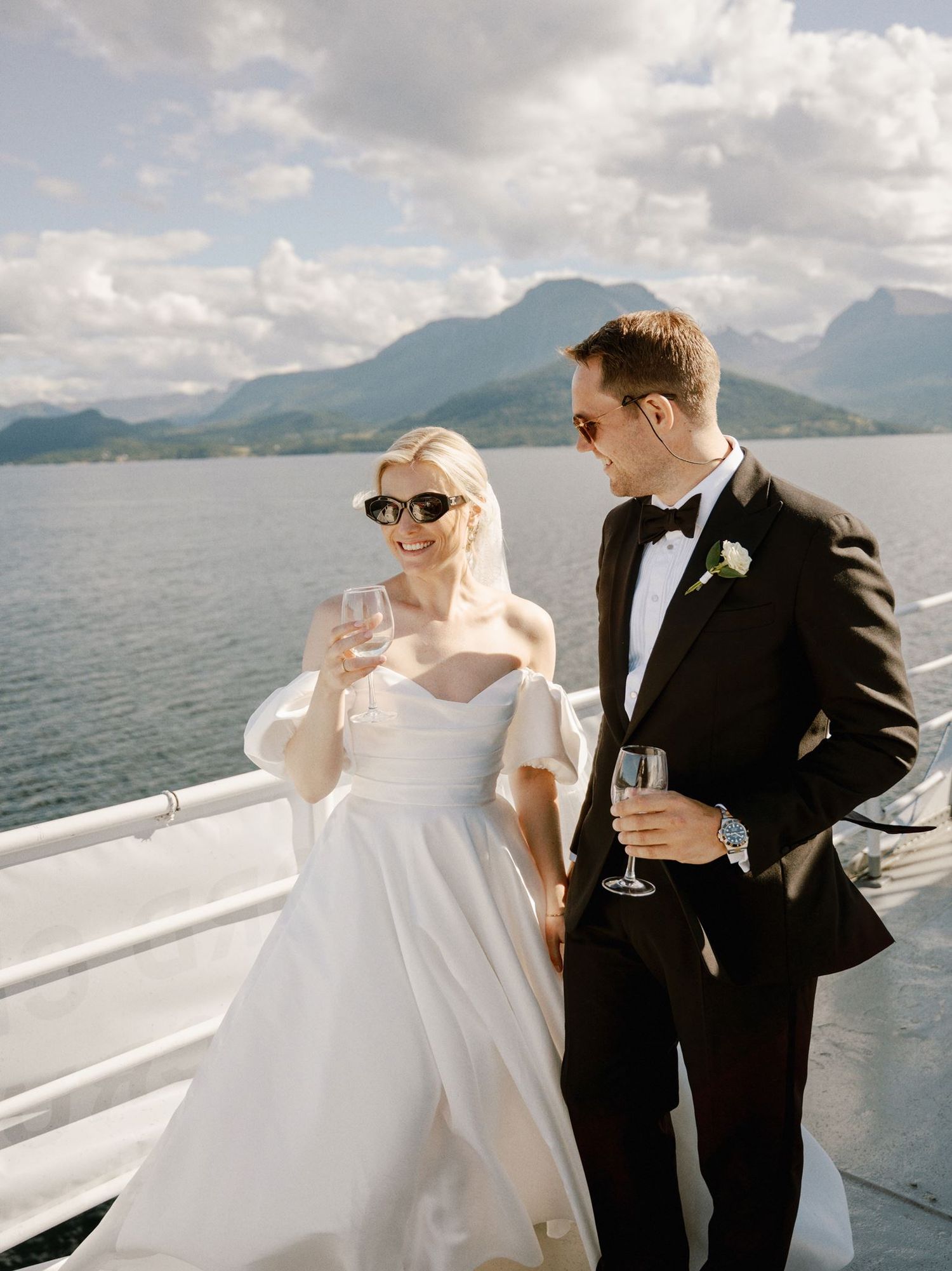 Bride in off-shoulder gown and groom in tuxedo toast with champagne on a boat deck with mountain backdrop.