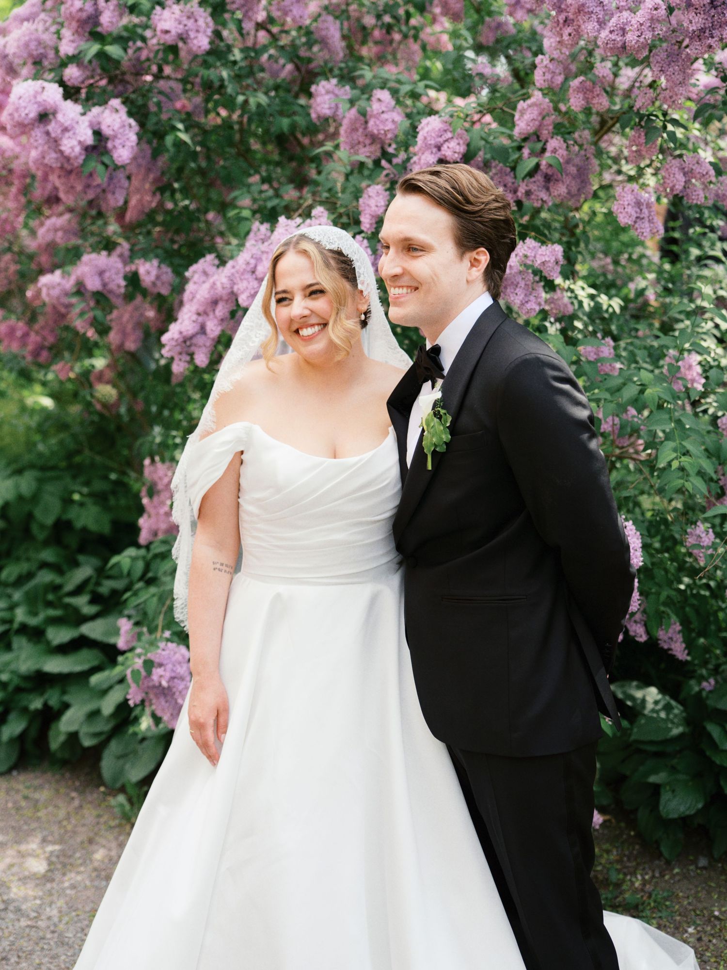 Bride and groom smiling together in front of blooming purple lilac bushes, bride in white gown and groom in black tuxedo.