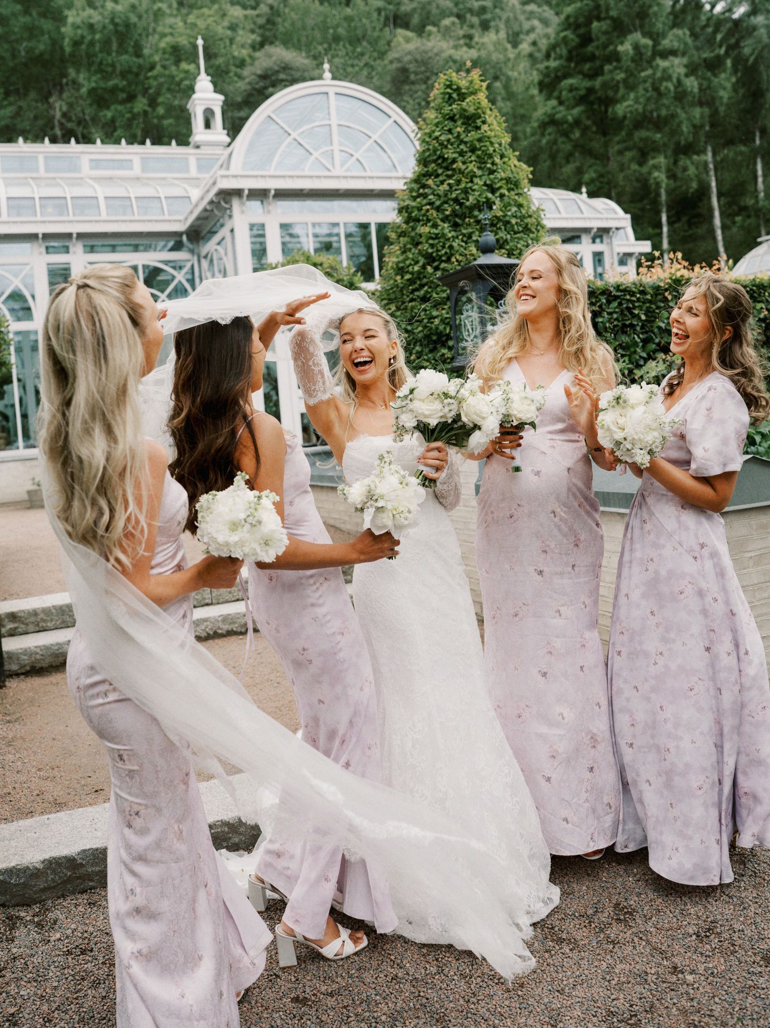 Bride in white gown laughs with bridesmaids in blush pink dresses holding white bouquets outside elegant greenhouse venue.