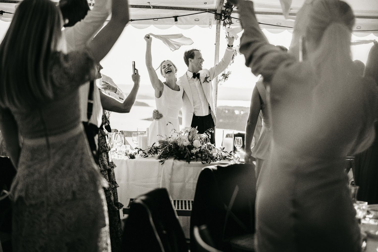 Black and white photo of joyful bride and groom celebrating at their wedding reception table.